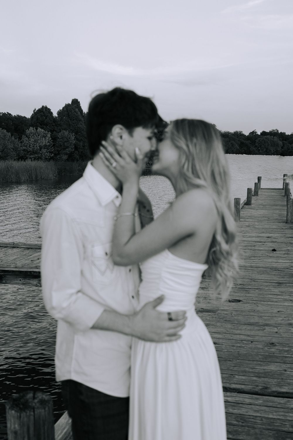 A romantic black and white photo series of a couple embracing on a wooden dock by the water.
