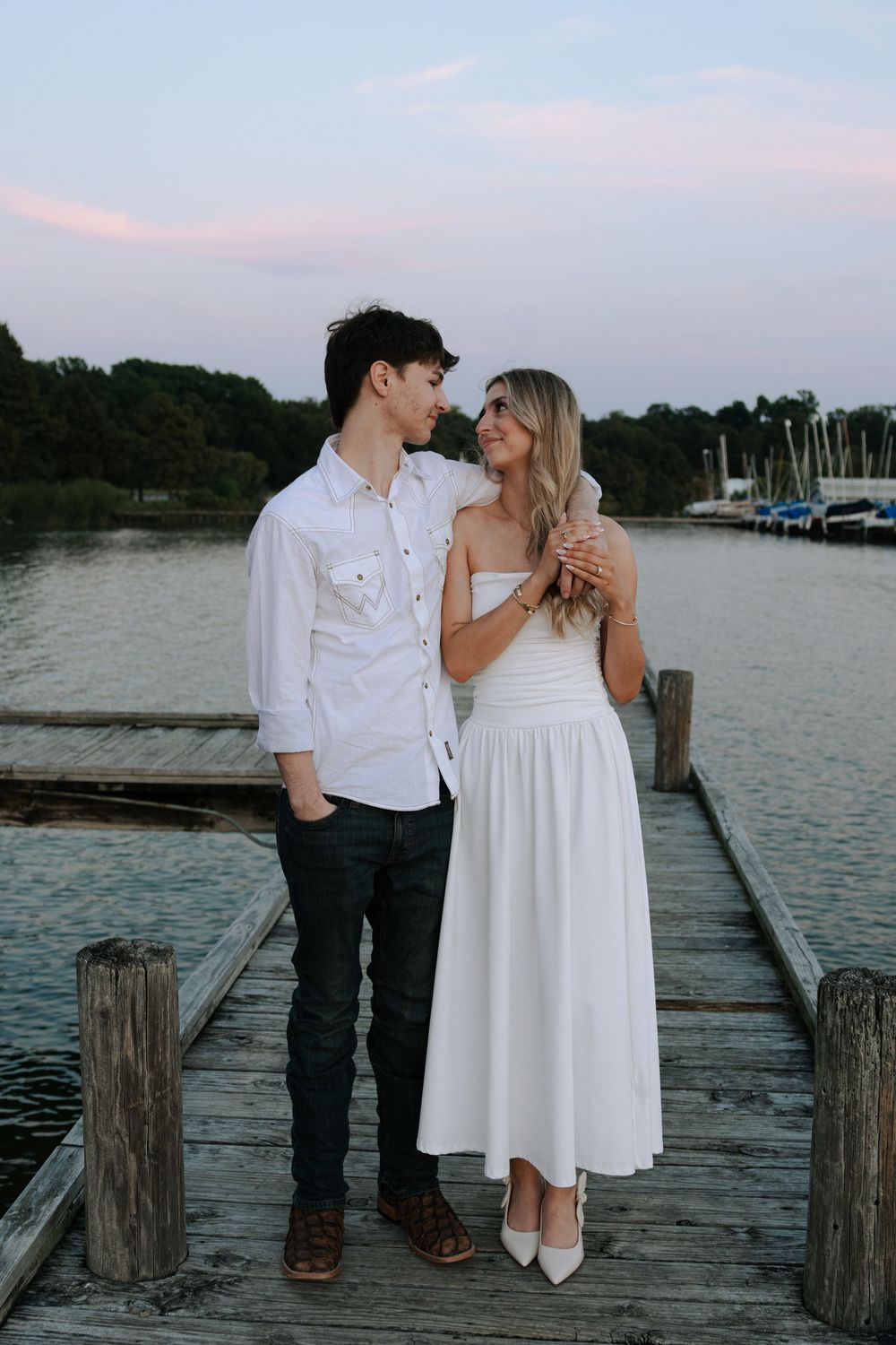 Romantic couple in white outfits share tender moments on a wooden dock by a lake at sunset.