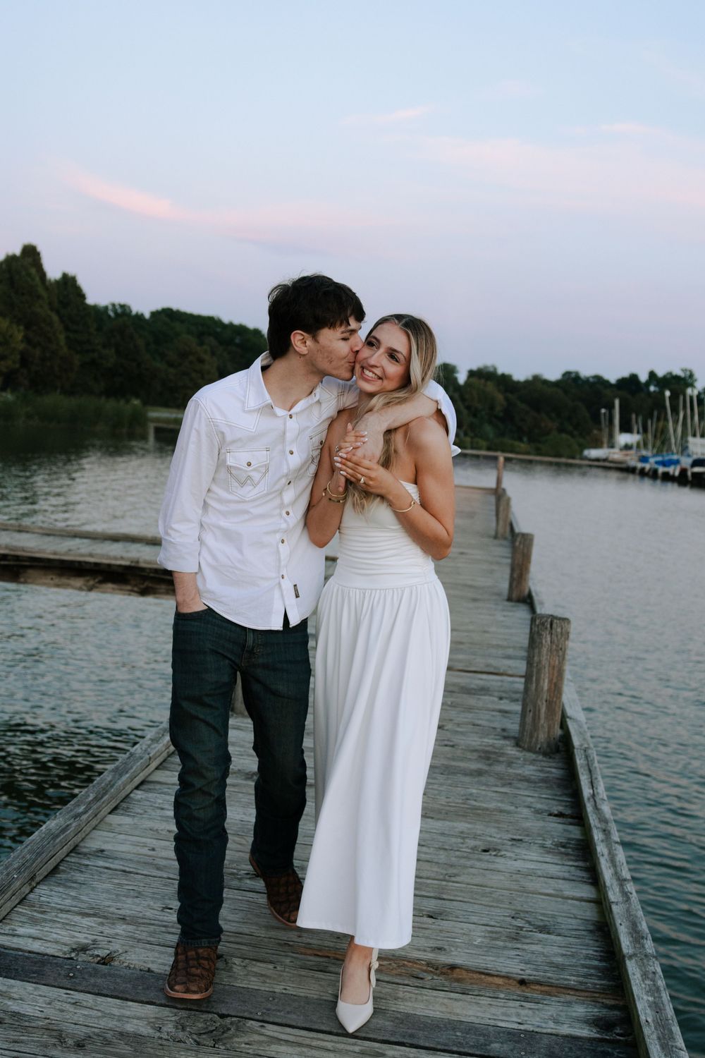 Sweet couple in coordinating white and dark outfits embrace on a lakeside pier during dusk.