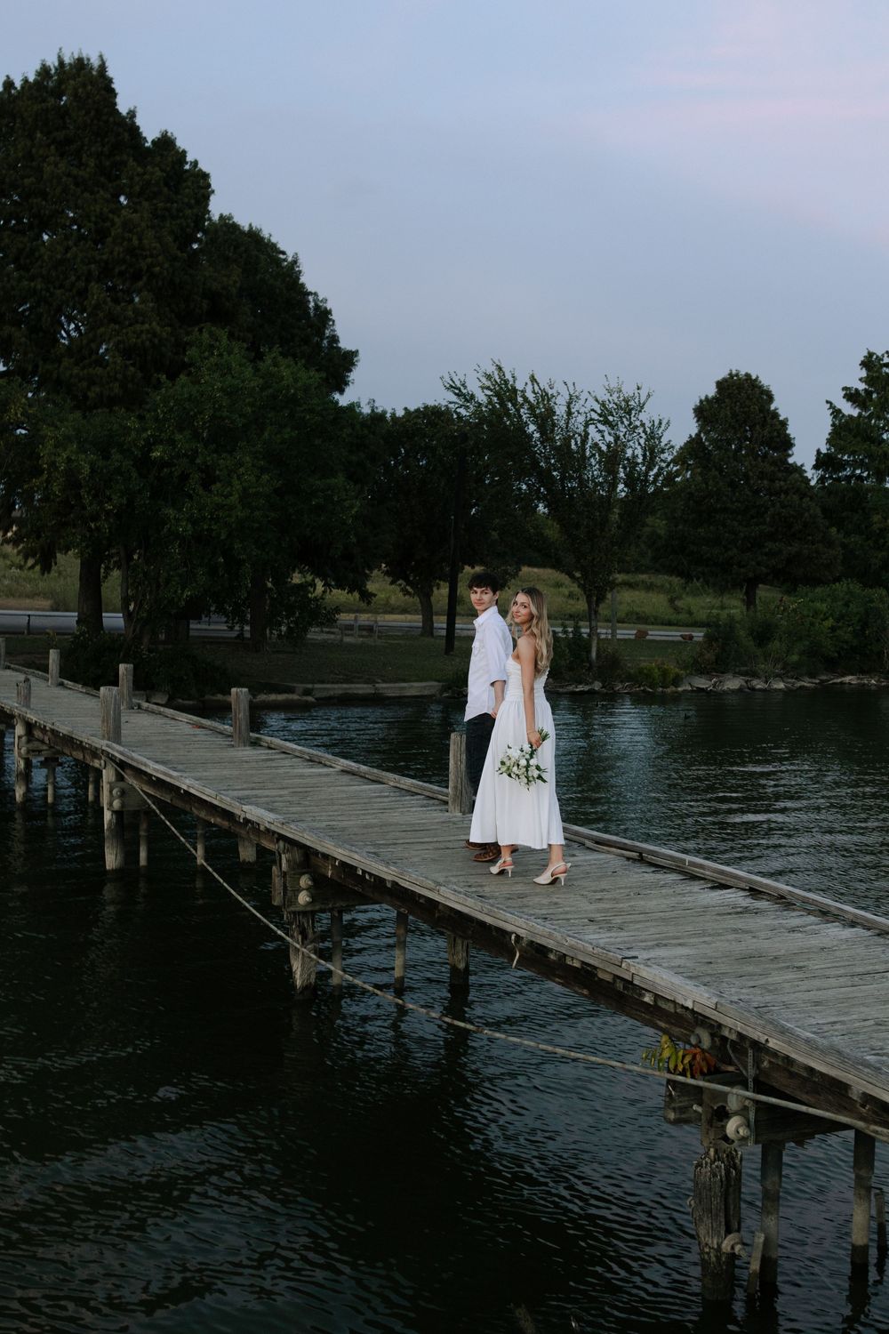 Two people walk along a weathered wooden pier extending into a tranquil lake at twilight.