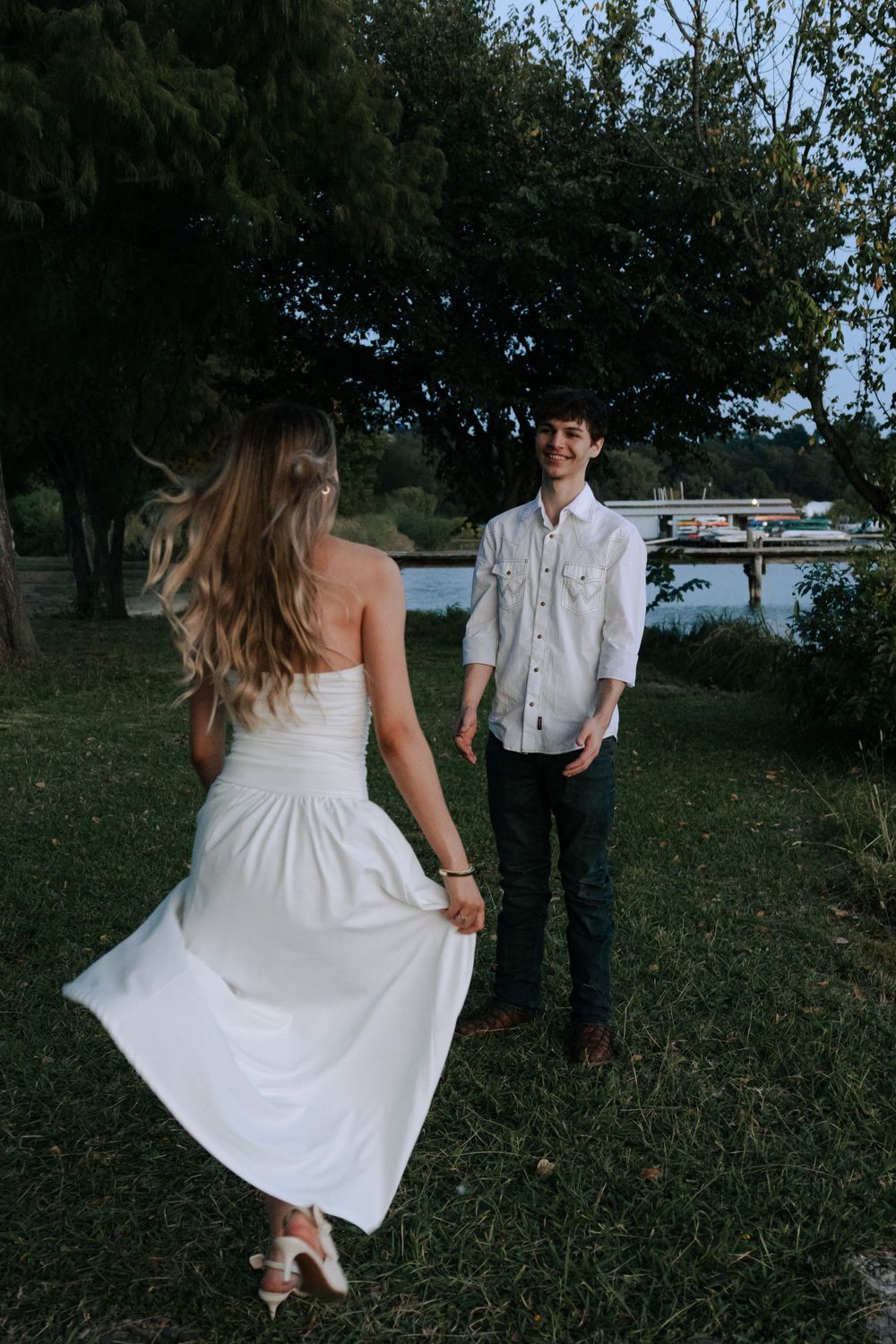 A couple in white attire poses playfully in a grassy outdoor setting near water.