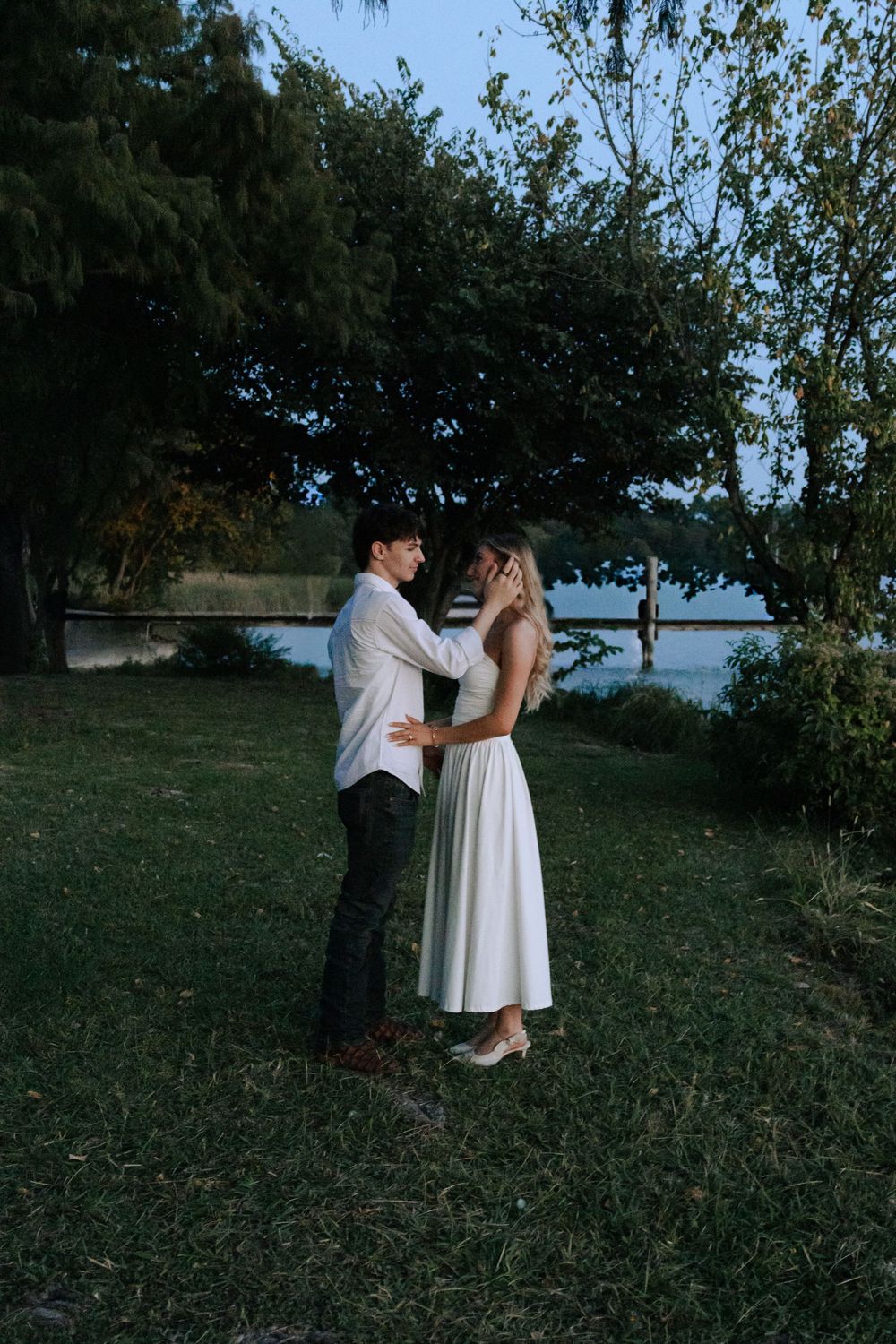 A couple dances together in a romantic outdoor setting with trees and grass at dusk.