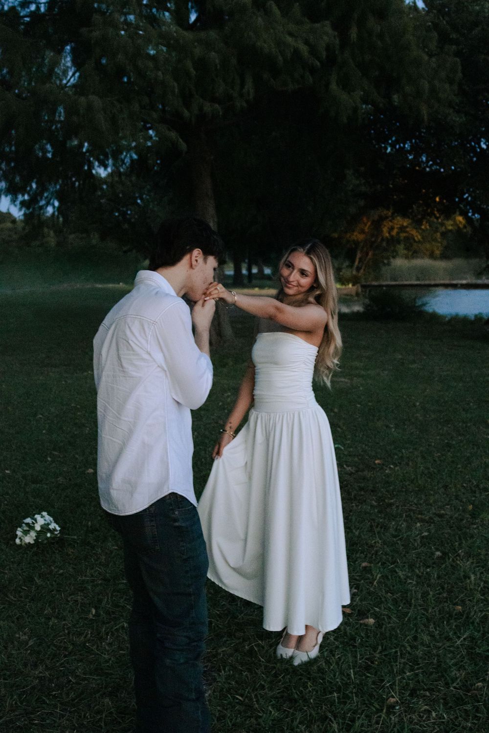 A romantic moment captured outdoors as a couple in white attire shares an intimate gesture by a lake at dusk.
