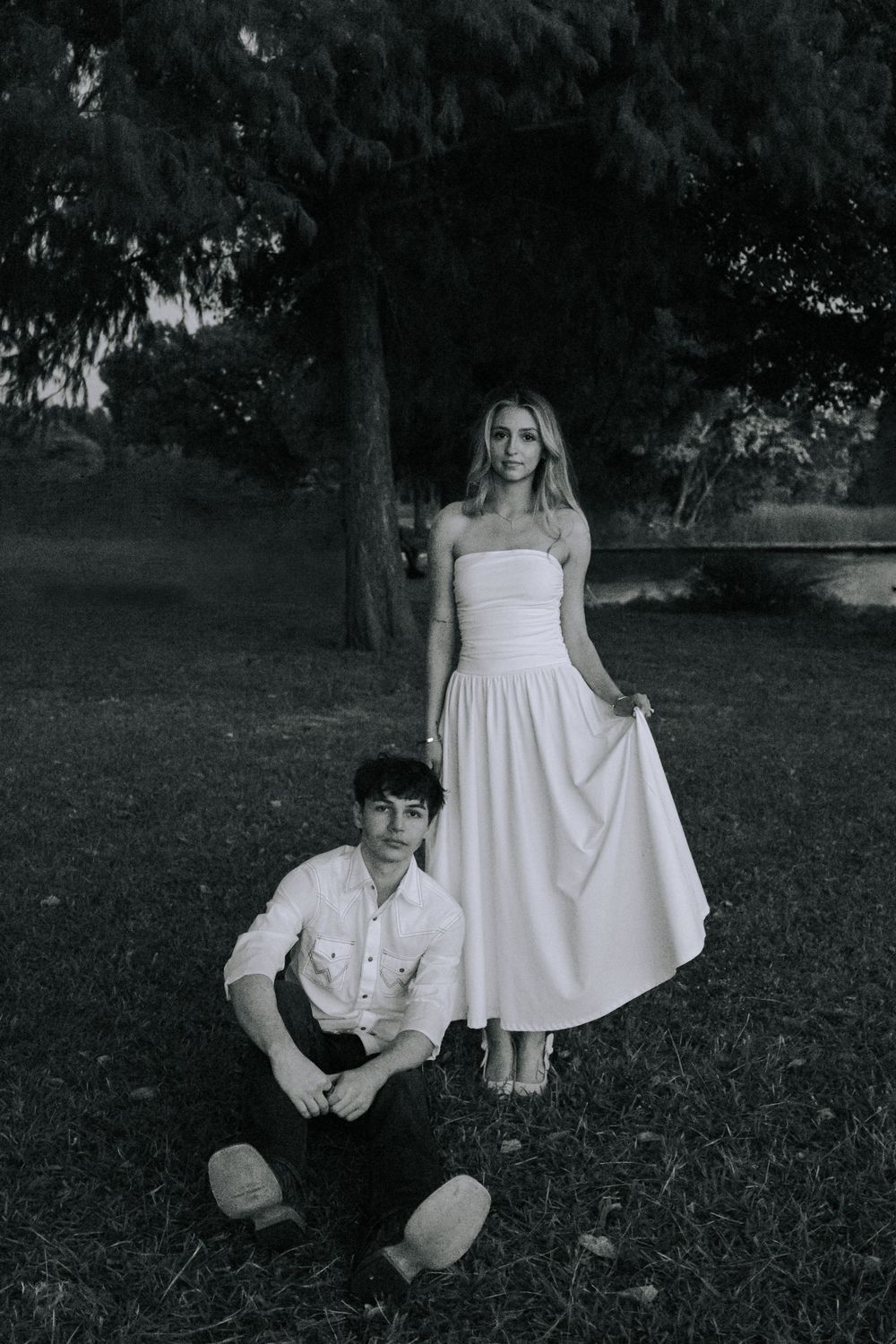 A couple in white clothing poses together on a grassy lawn with trees in the background.
