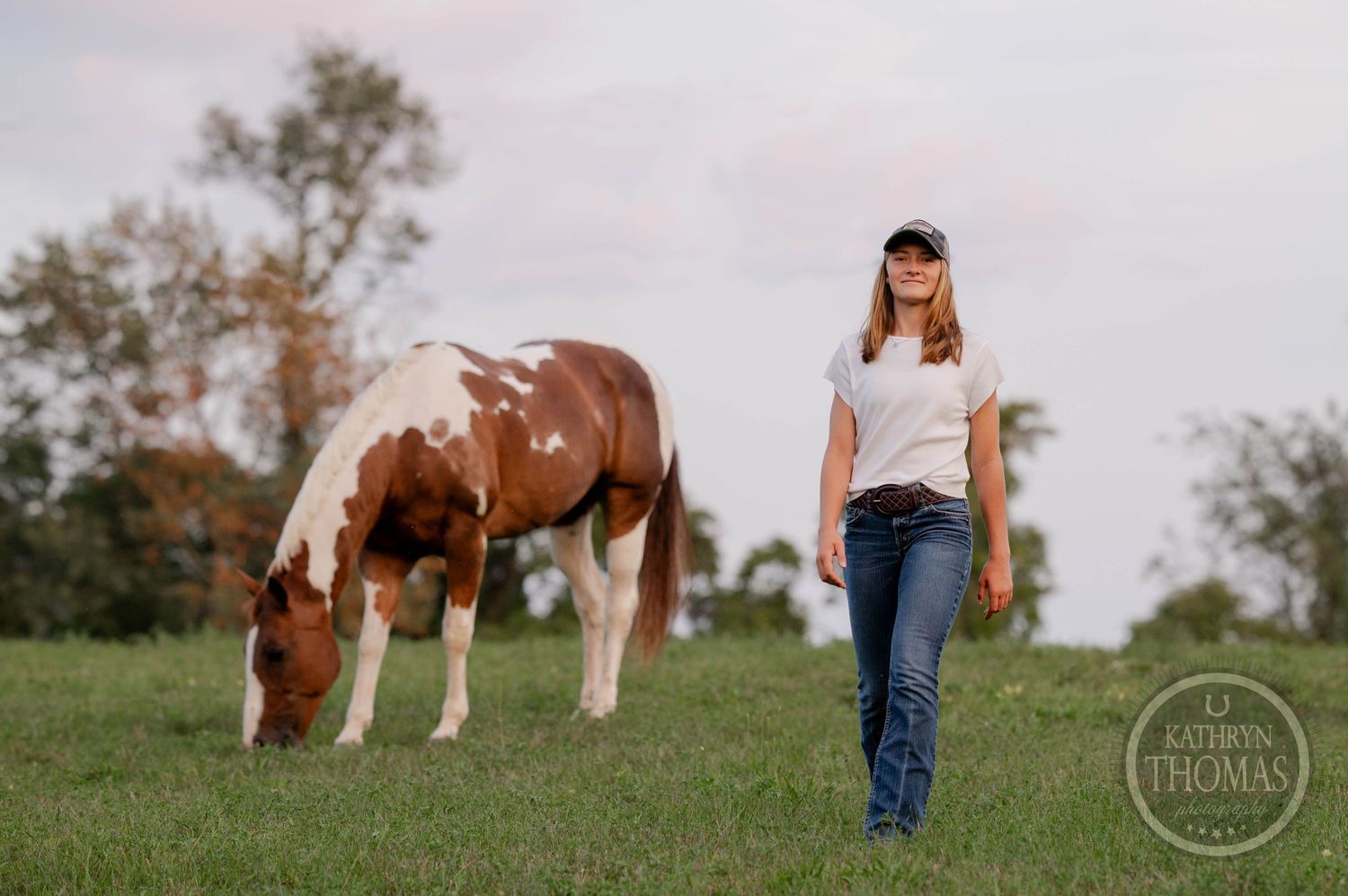 A brown and white paint horse grazes in a green pasture while someone stands nearby in casual attire.