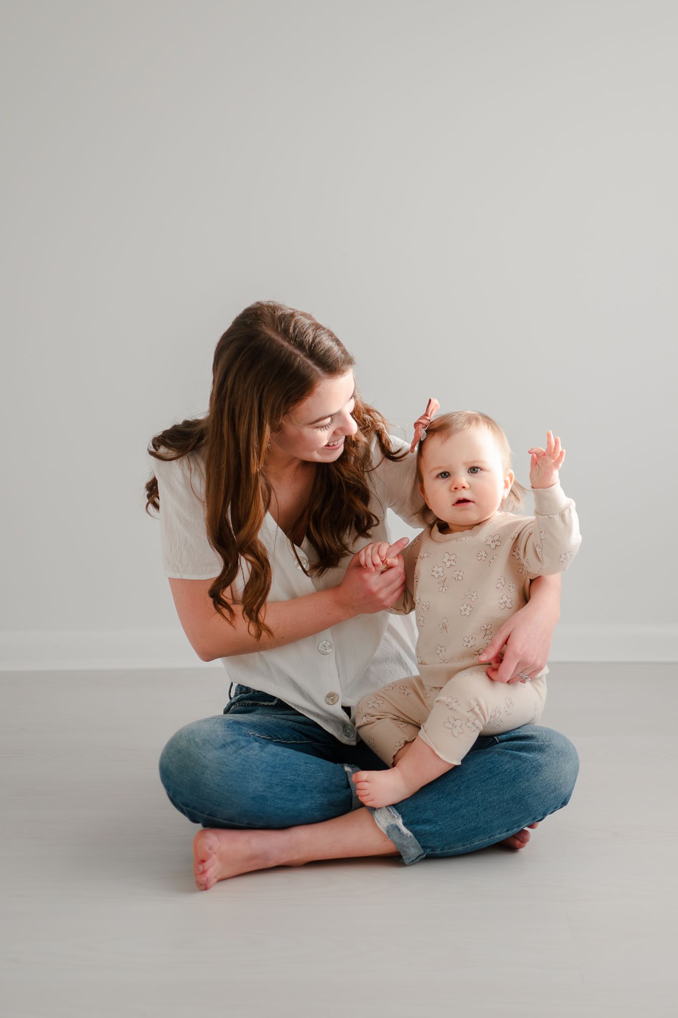 Tender parenting moment captured as mom and baby play together while sitting on floor against light gray backdrop.