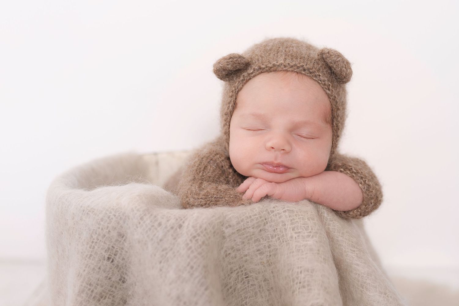 Sleeping newborn wearing brown teddy bear outfit resting on soft blanket during professional photo session.