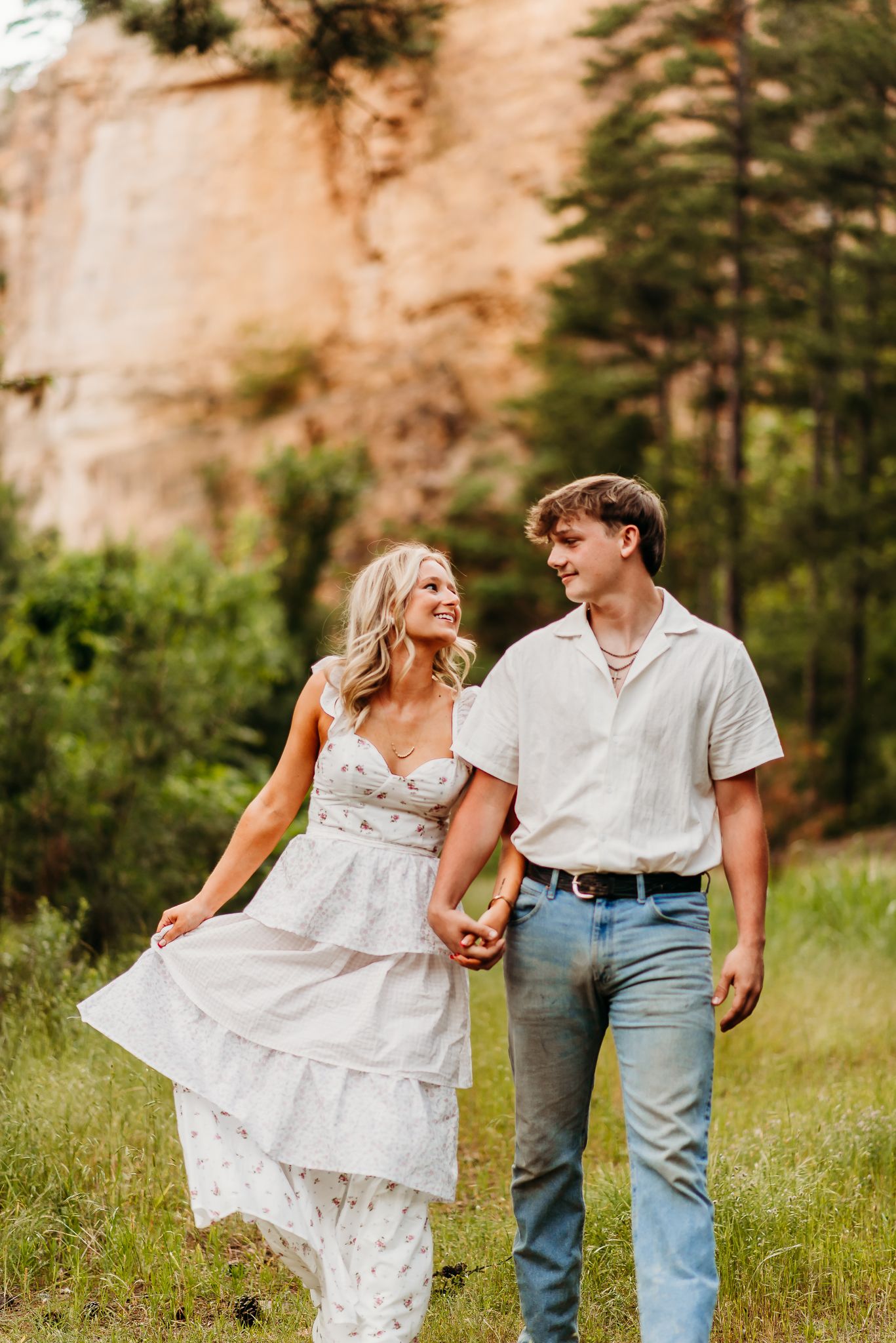 Couple walking hand in hand in white dress and casual attire against dramatic cliff backdrop.