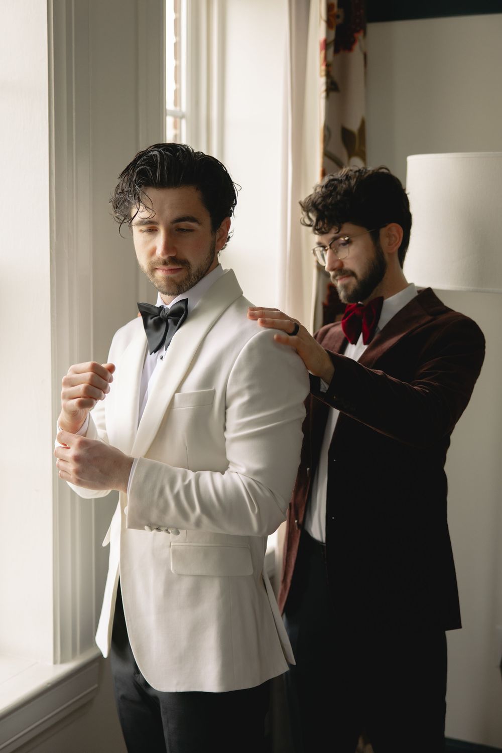 Groomsman in burgundy velvet suit helping groom in white tuxedo jacket prepare near bright window.