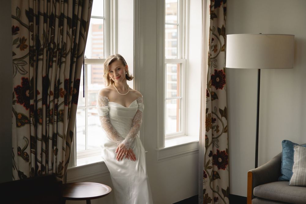 Elegant bride in an off-shoulder white lace wedding gown smiling by a sunlit window with floral curtains.
