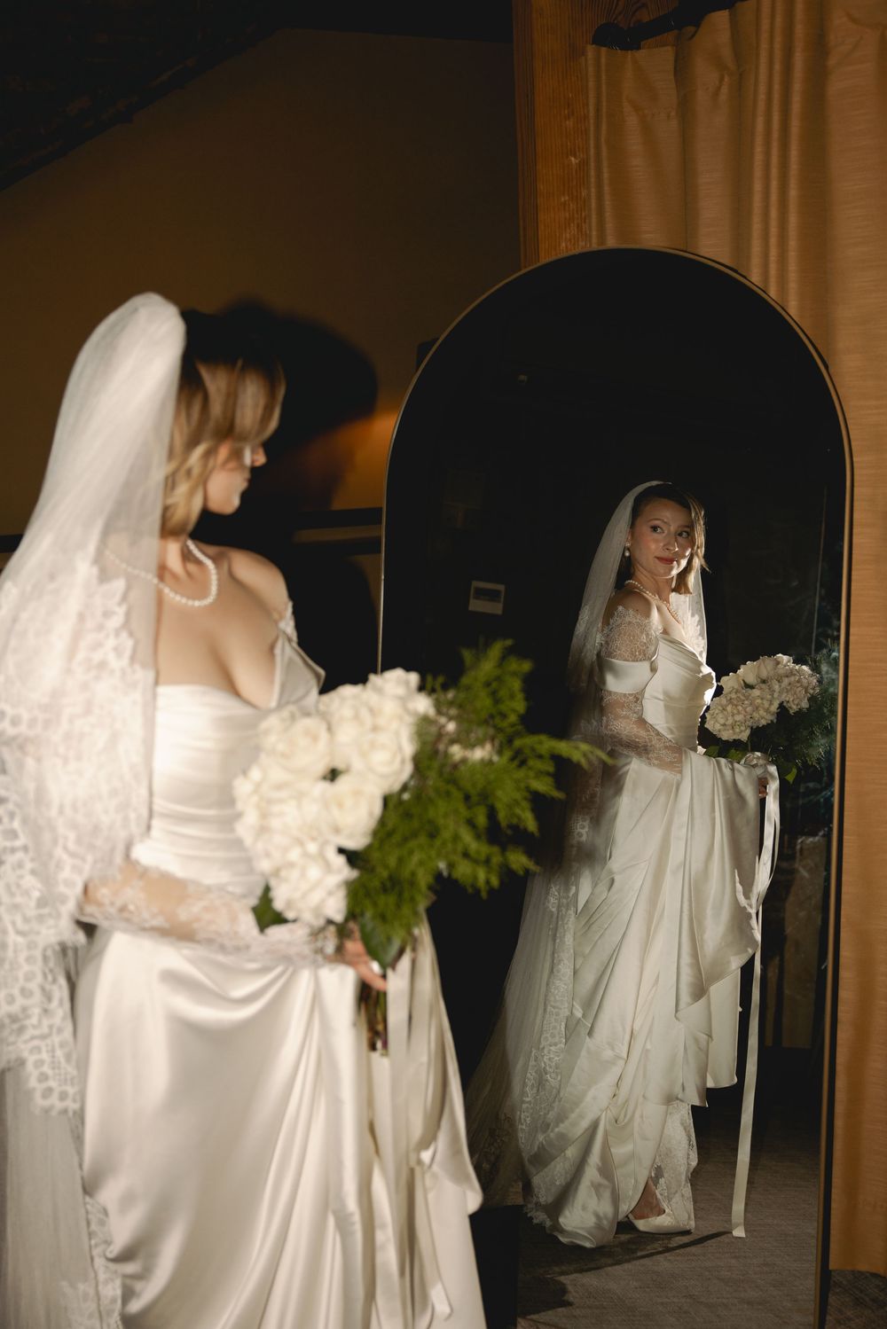 Blonde bride in white satin gown with lace veil holding white floral bouquet reflected in arched mirror.