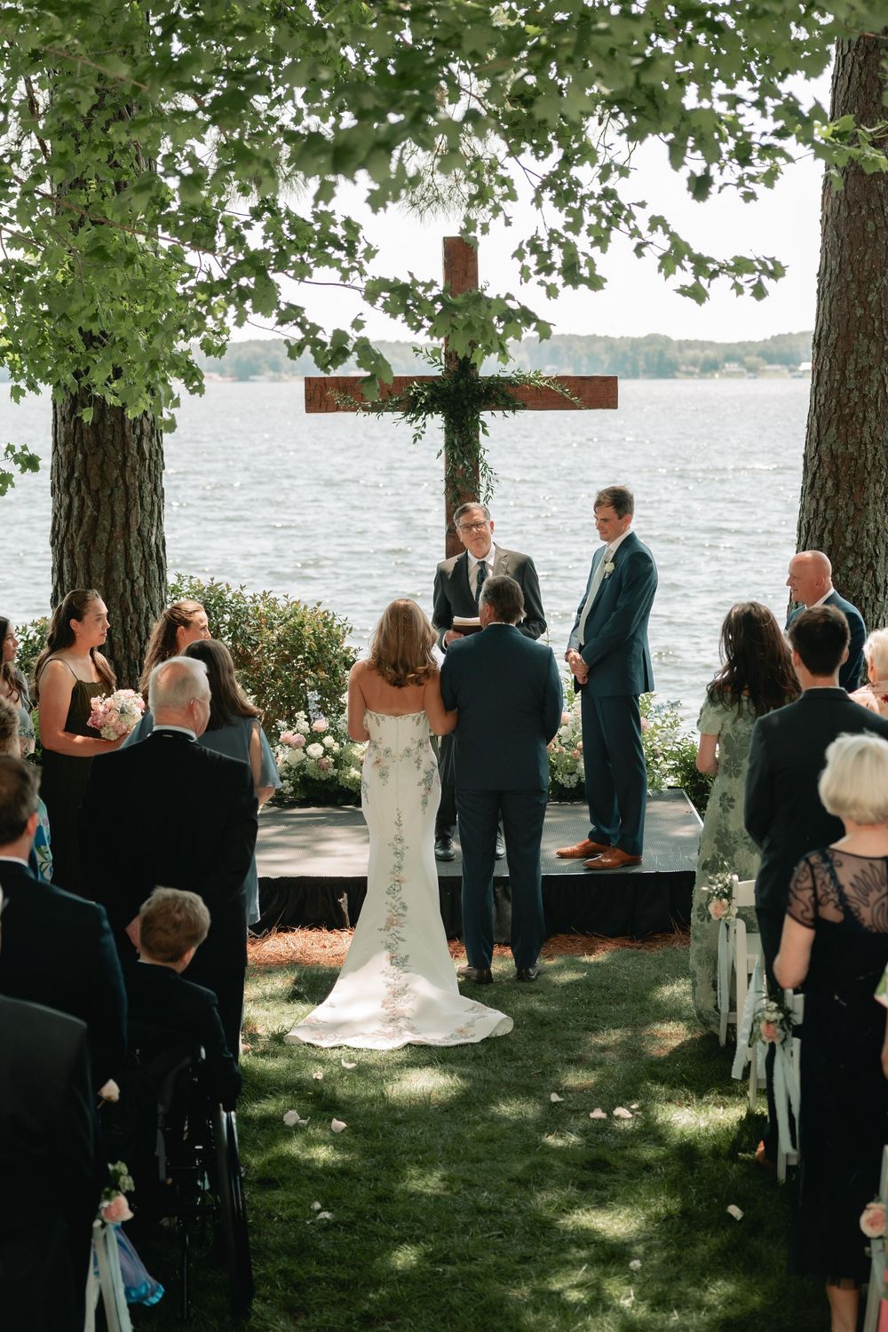 Bride and groom exchanging vows at lakeside outdoor ceremony beneath wooden cross and tree canopy.