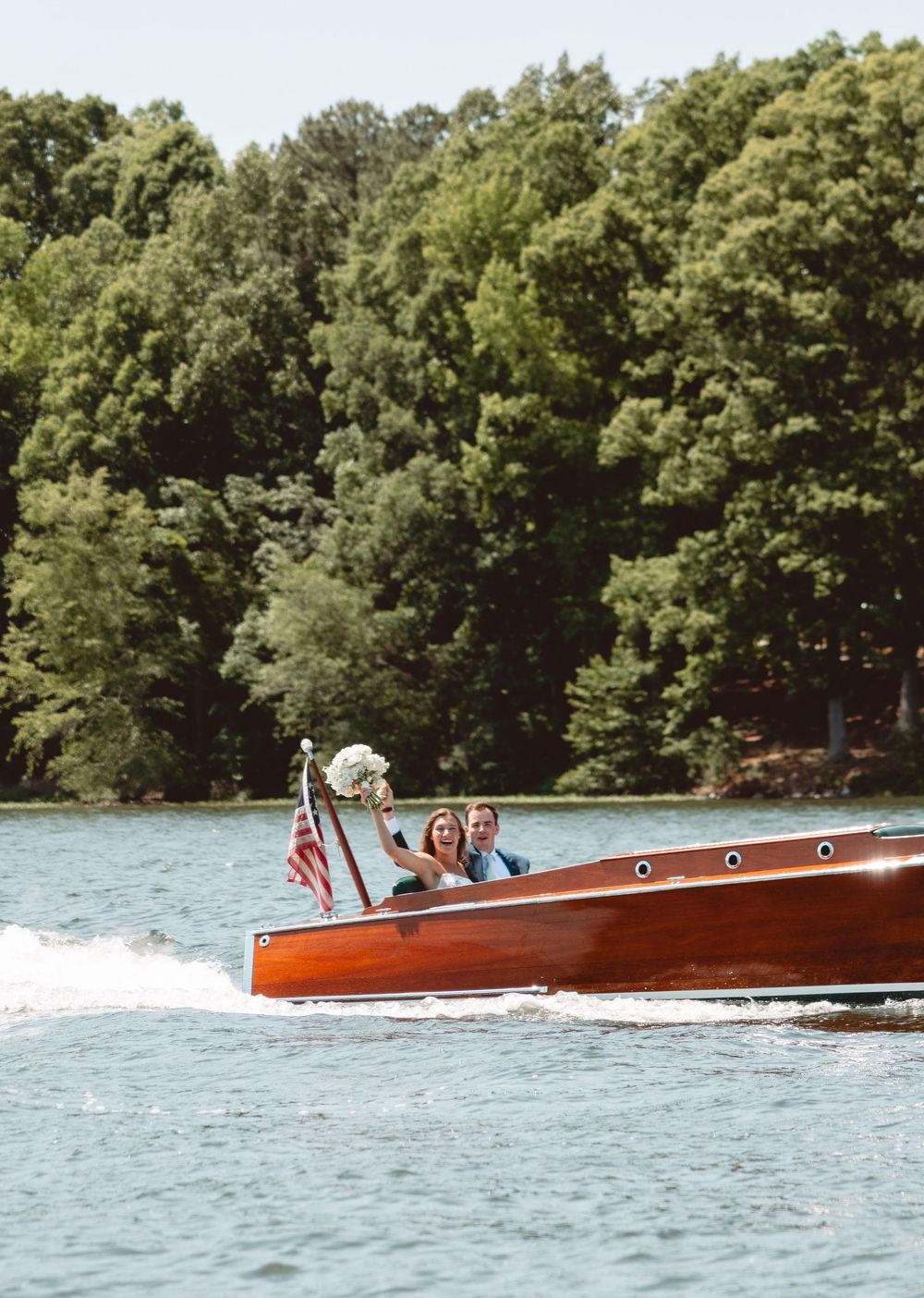 Bride and groom ride classic wooden speedboat across sparkling lake after wedding ceremony.
