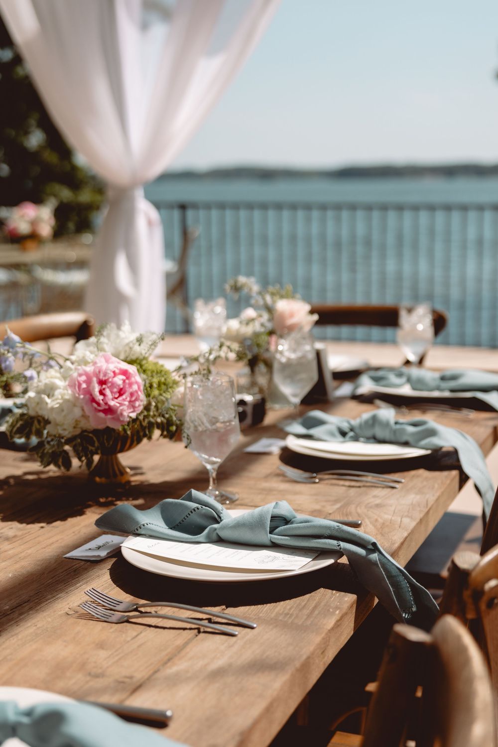 Elegant outdoor lakeside wedding reception table set with sage napkins and pink peony centerpieces.