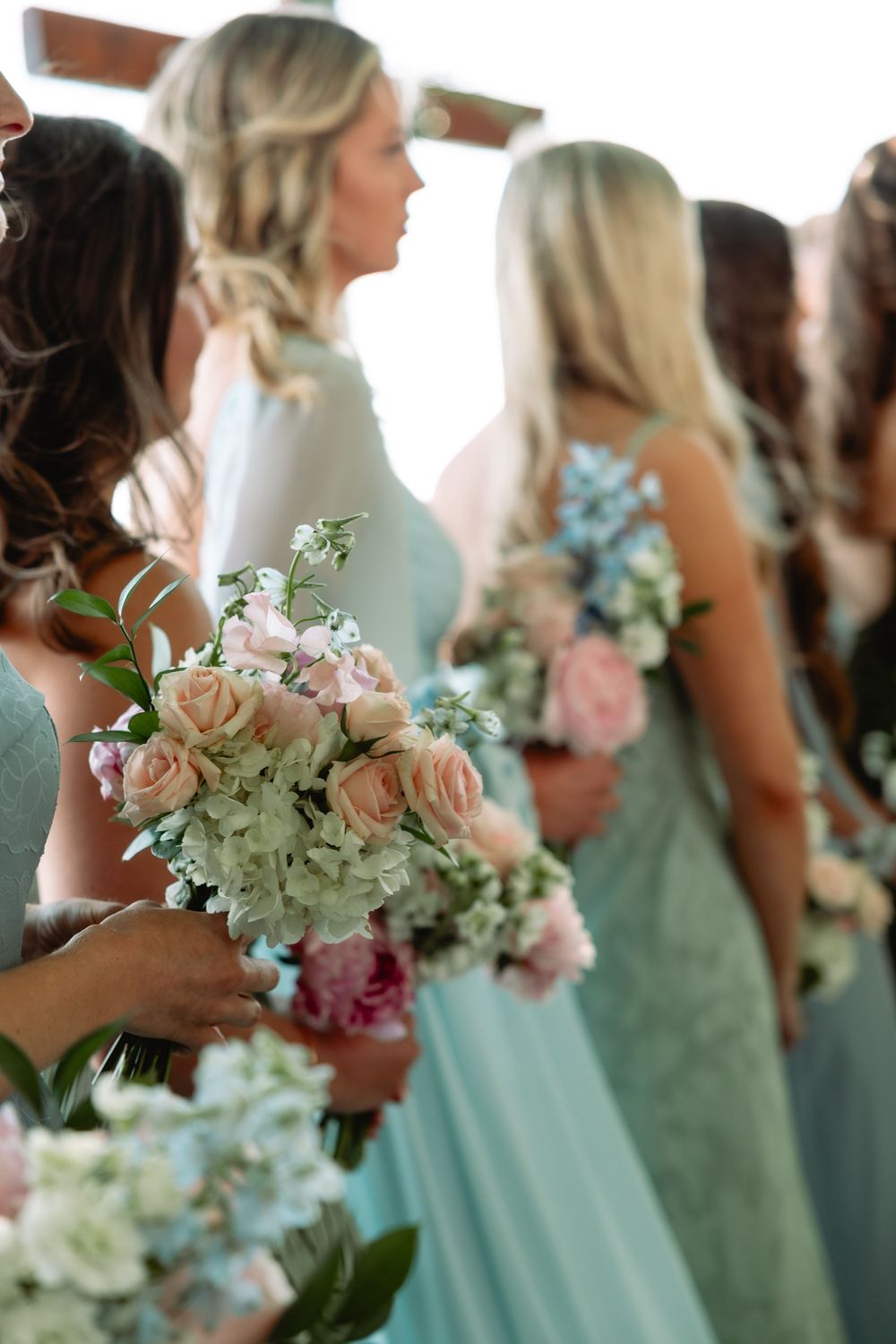Bridesmaids in soft blue gowns holding bouquets of pink roses and hydrangeas at wedding ceremony.