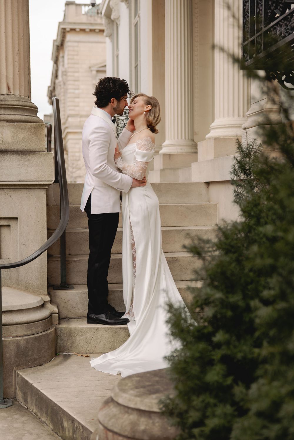 Bride and groom sharing a romantic kiss on stone steps of a grand building with tall columns.