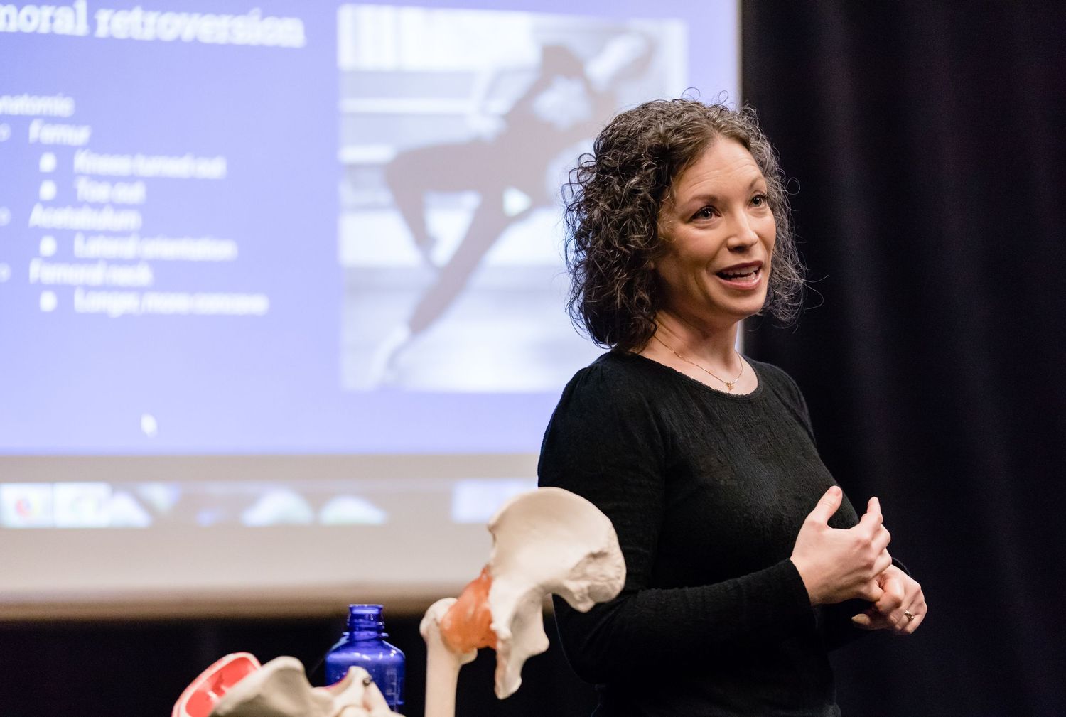 Speaker in black top presents at a conference with projection screen and anatomical models visible in background.