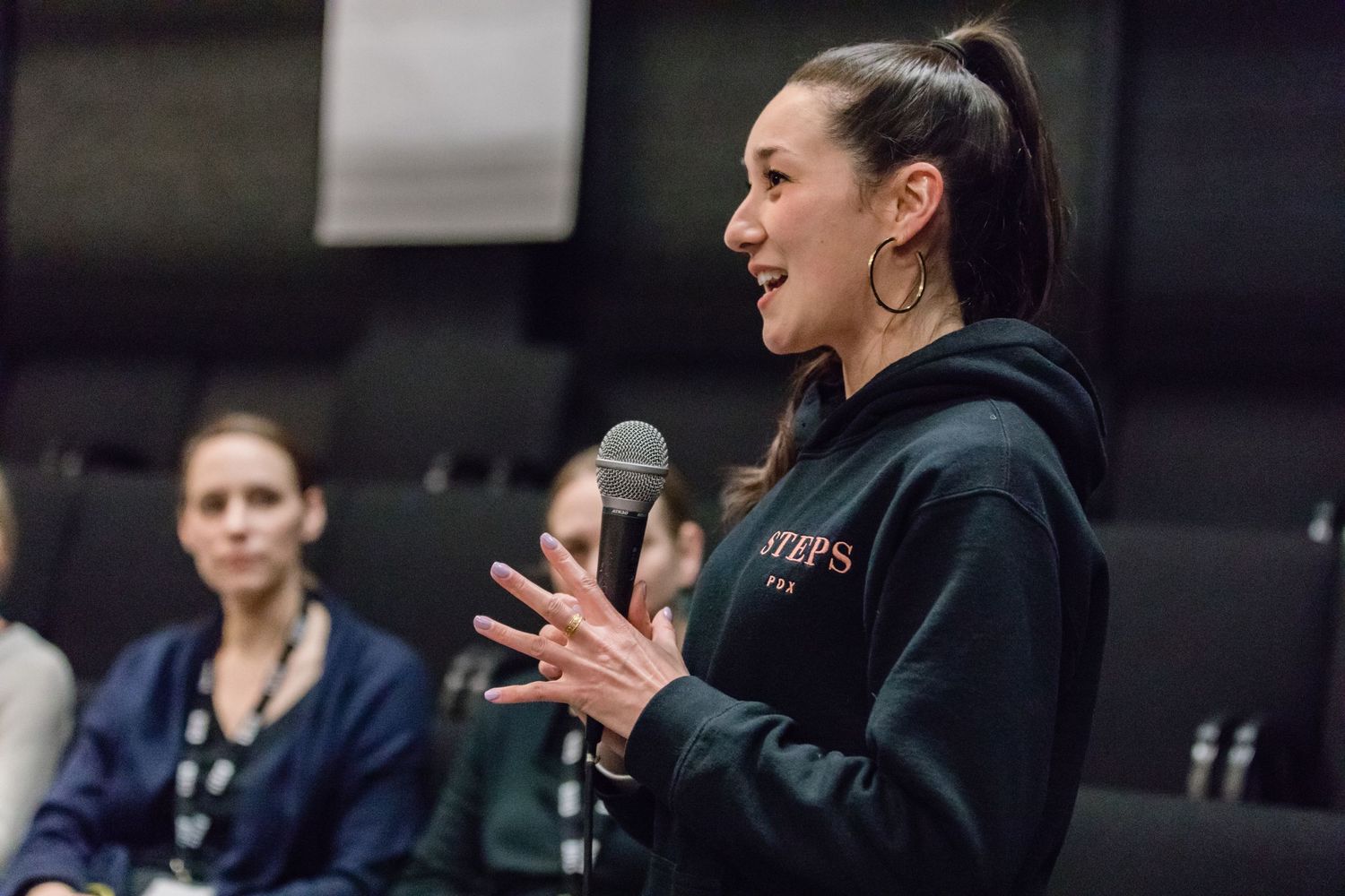 A speaker holds a microphone while gesturing animatedly during a presentation, wearing a dark hoodie.