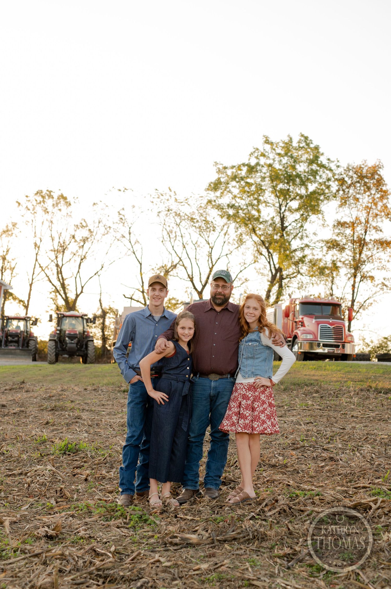 A casual outdoor family portrait taken at sunset with farm equipment and trees in the background.