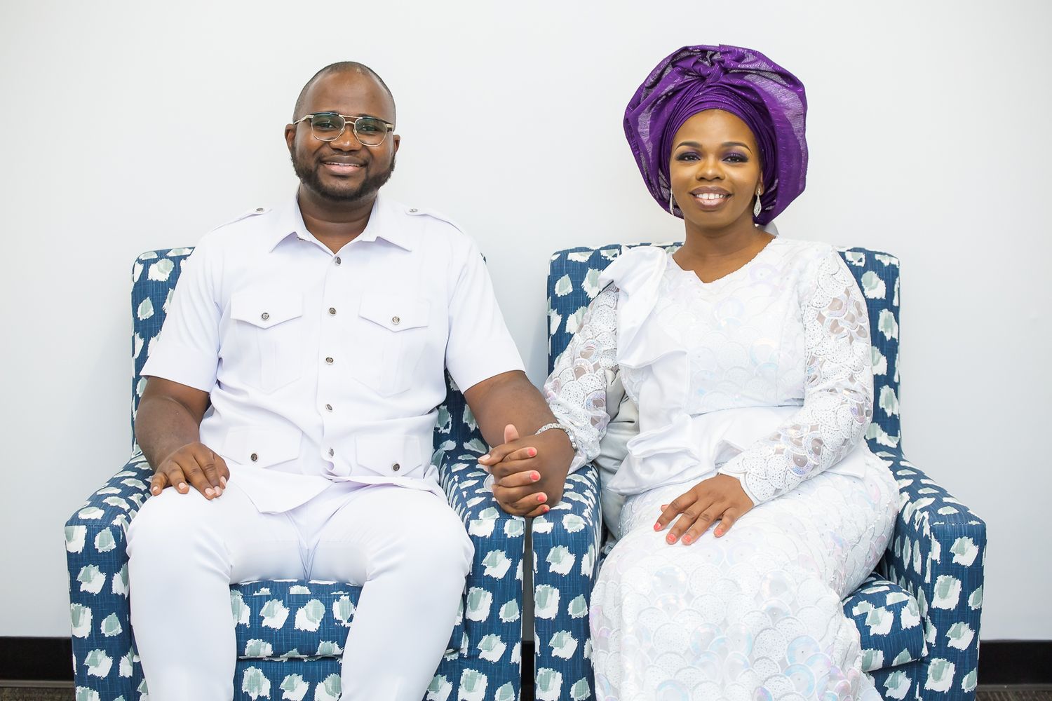 A couple dressed in white traditional attire sit together holding hands on a blue and white patterned couch.