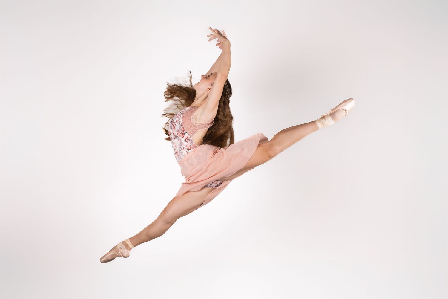 A dancer in a flowing pink dress performs a graceful leap against a white background.