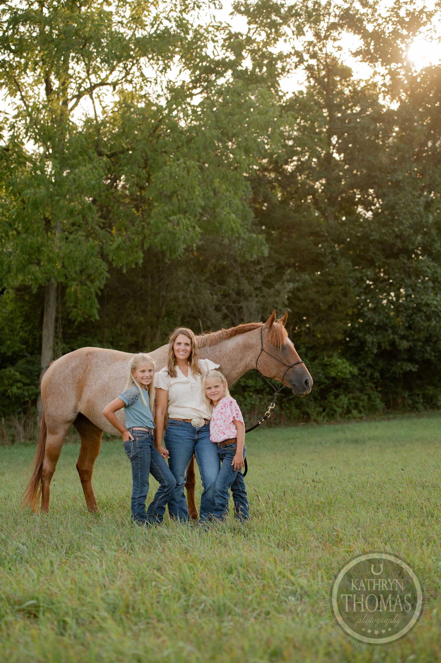 Three people pose together with their brown horse against a backdrop of trees at dusk.