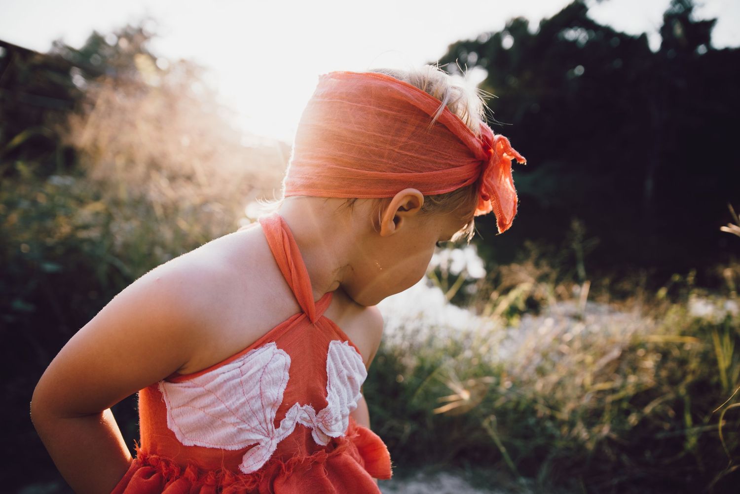A person in a coral orange outfit and matching headband stands in a grassy field at sunset.
