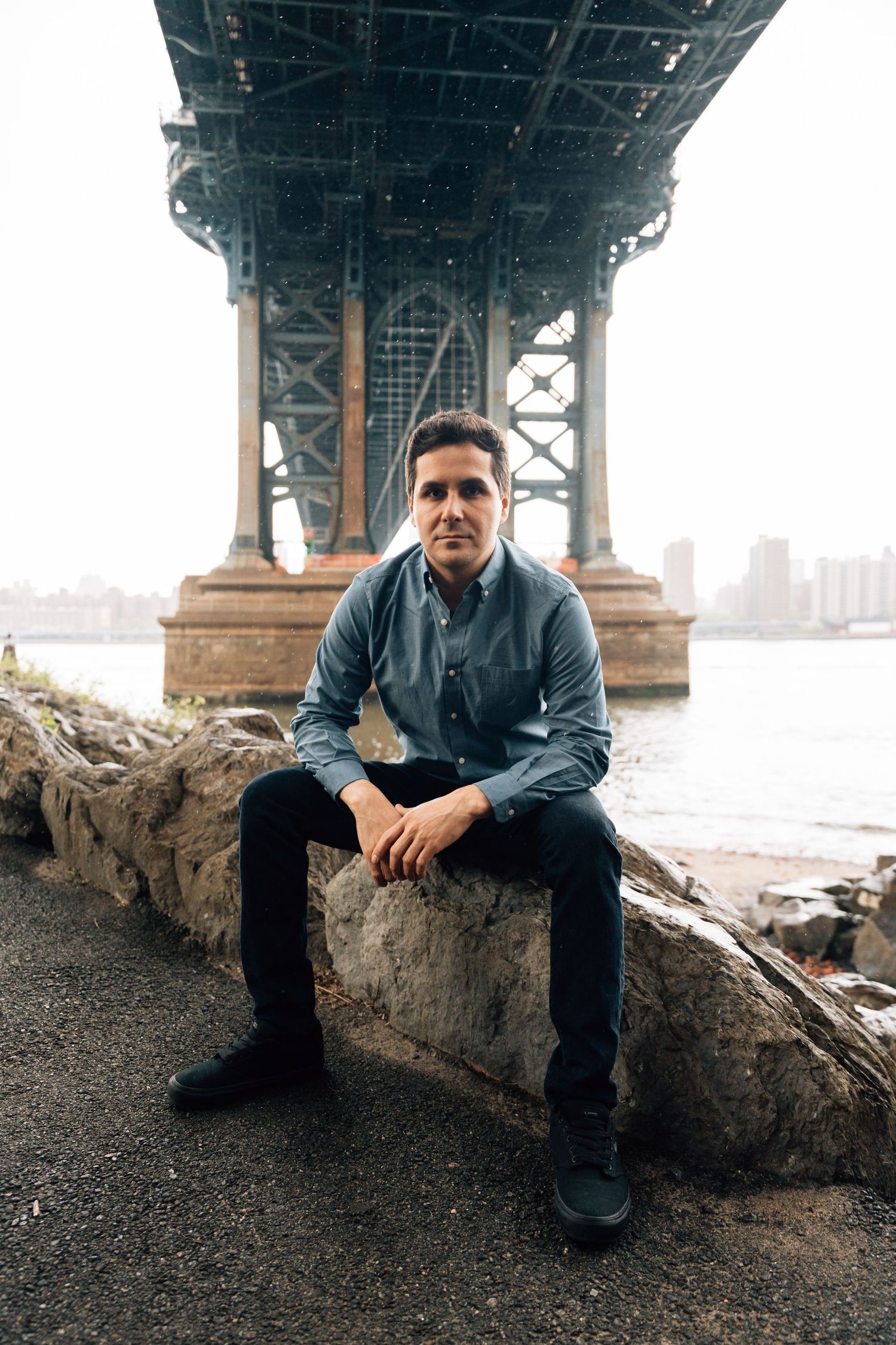 Person sits on stone wall beneath Manhattan Bridge with East River and cityscape in view.