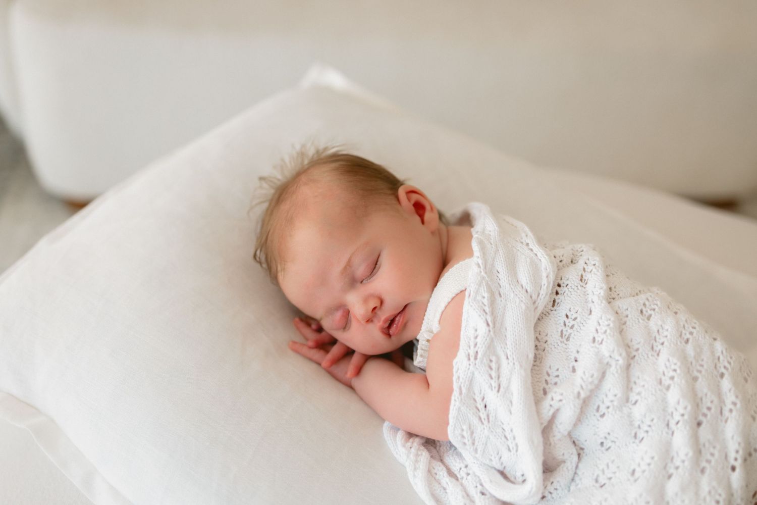 Baby wearing white knitted outfit lying on soft white bedding with gentle natural lighting.