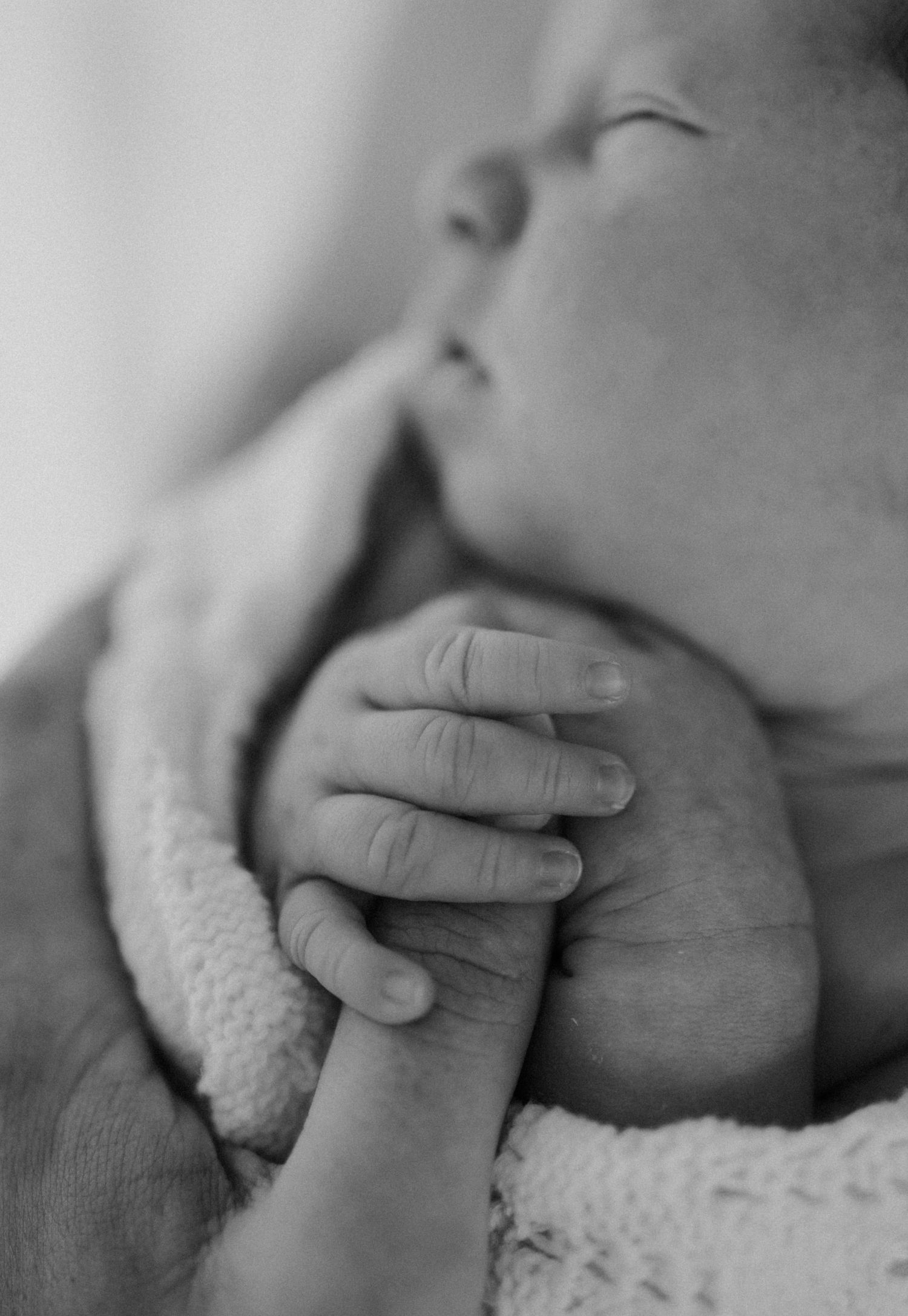 A tender moment captured between a mother and sleeping newborn baby wrapped in white blanket in warm lighting.