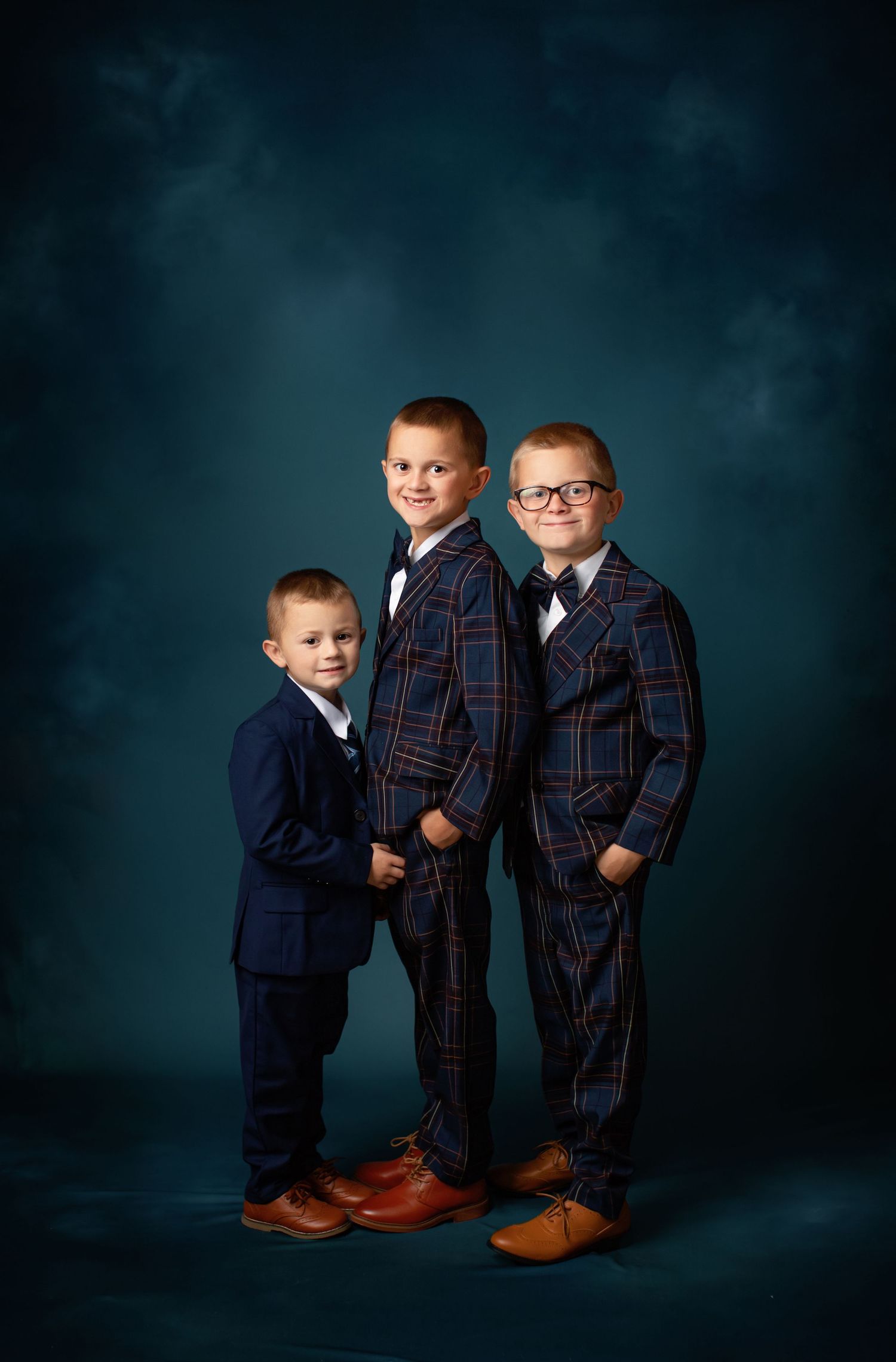 Three young children pose together in formal plaid and navy suits against a dark teal studio backdrop.