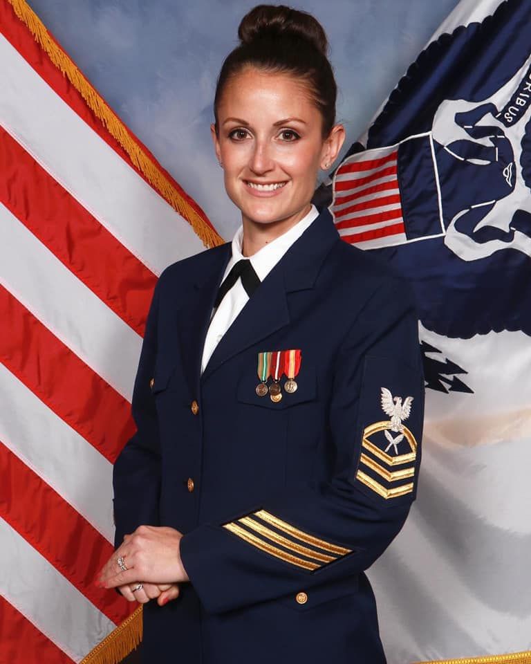 A service member in US Coast Guard dress blue uniform poses with American flag and Coast Guard flag in the background.