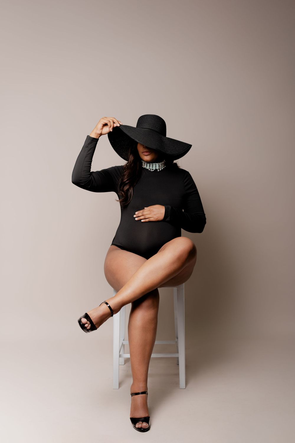Stylish maternity portrait with woman in black bodysuit and wide-brim hat sitting on white stool.