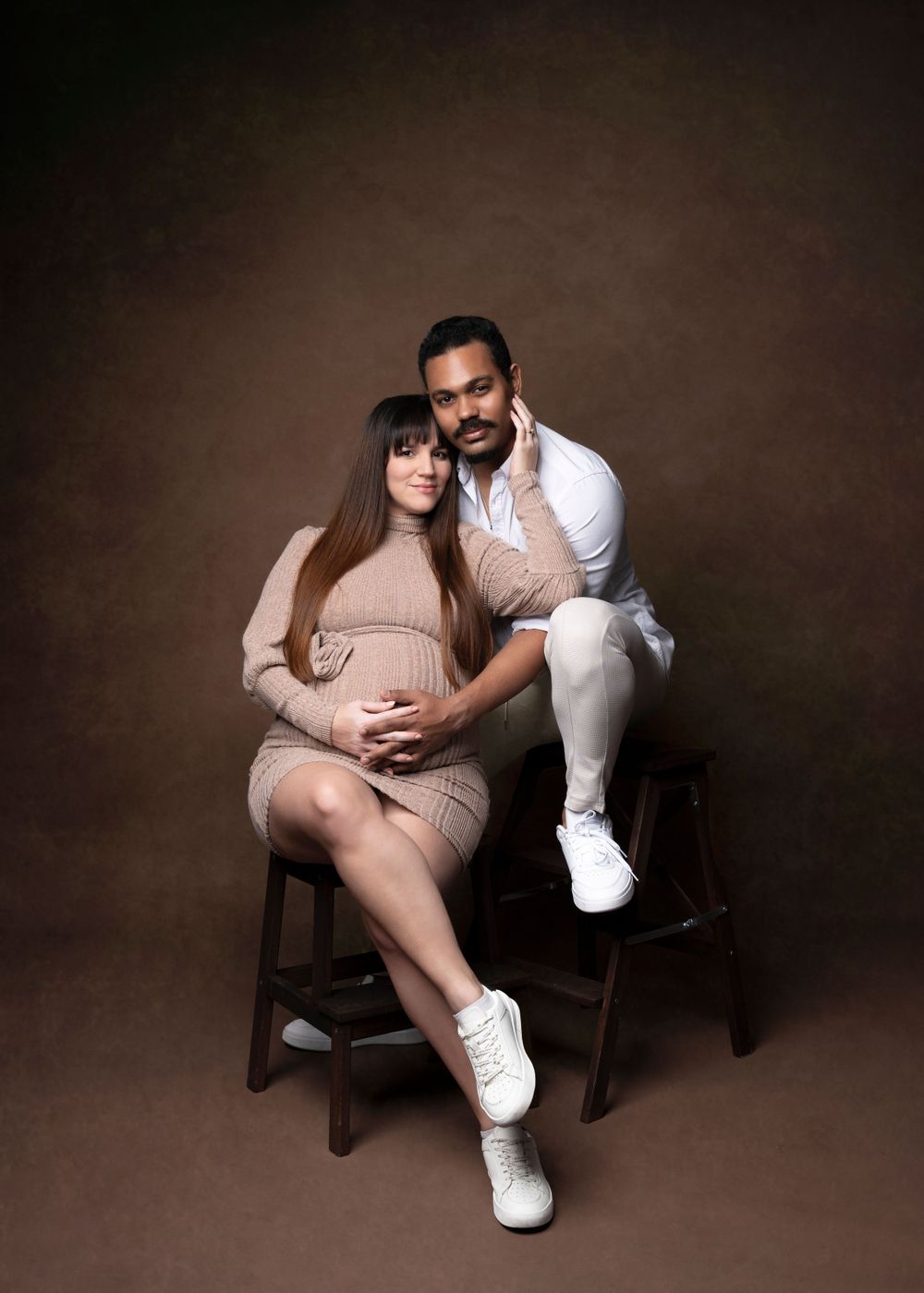 Intimate maternity studio portrait of couple in neutral tones against brown background.