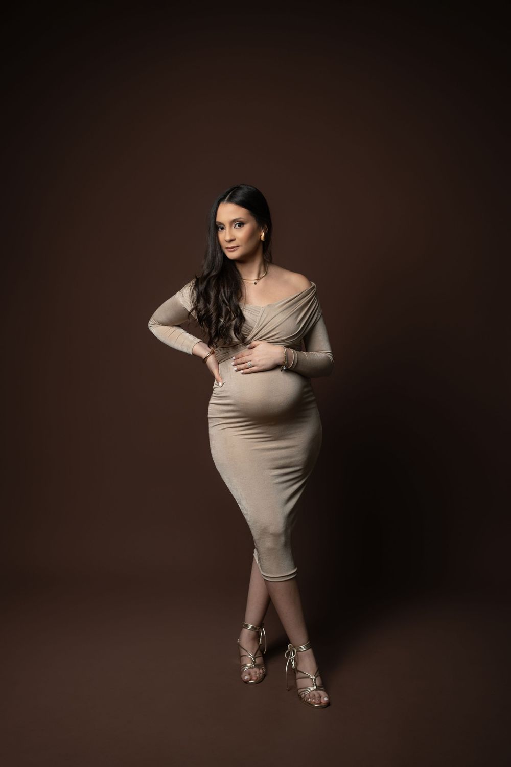 Sophisticated maternity pose in fitted beige dress against chocolate brown studio background.