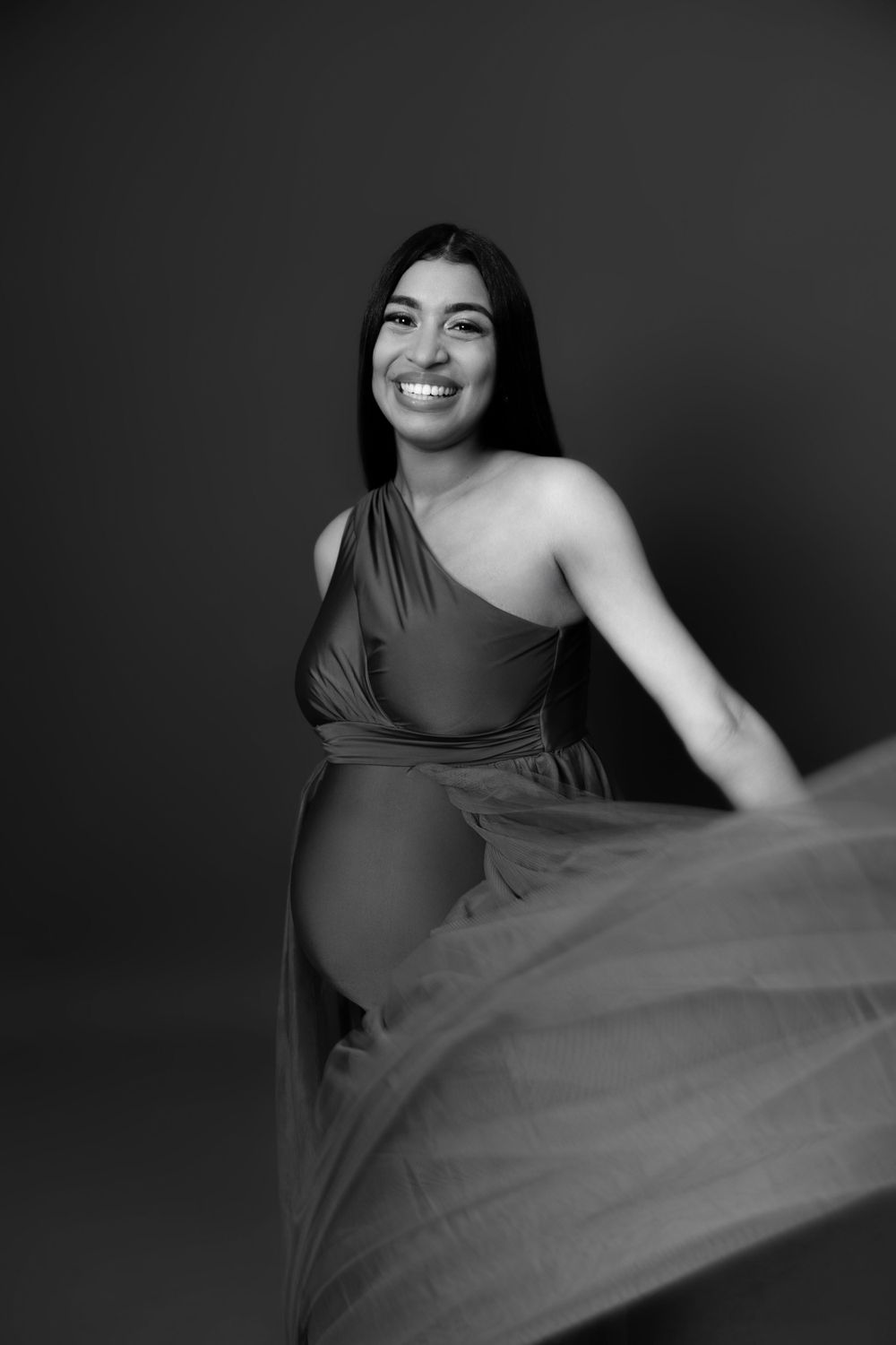 Elegant maternity photoshoot featuring a one-shoulder flowing dress against a dark background.