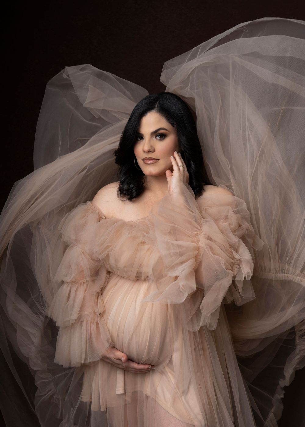 Dreamy maternity portrait in flowing peach tulle gown against dark backdrop.