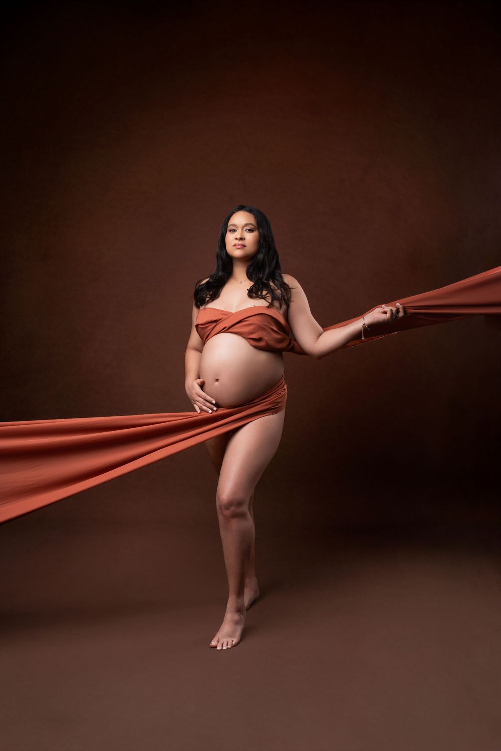 Artistic maternity portrait with flowing terracotta fabric against dark brown background.