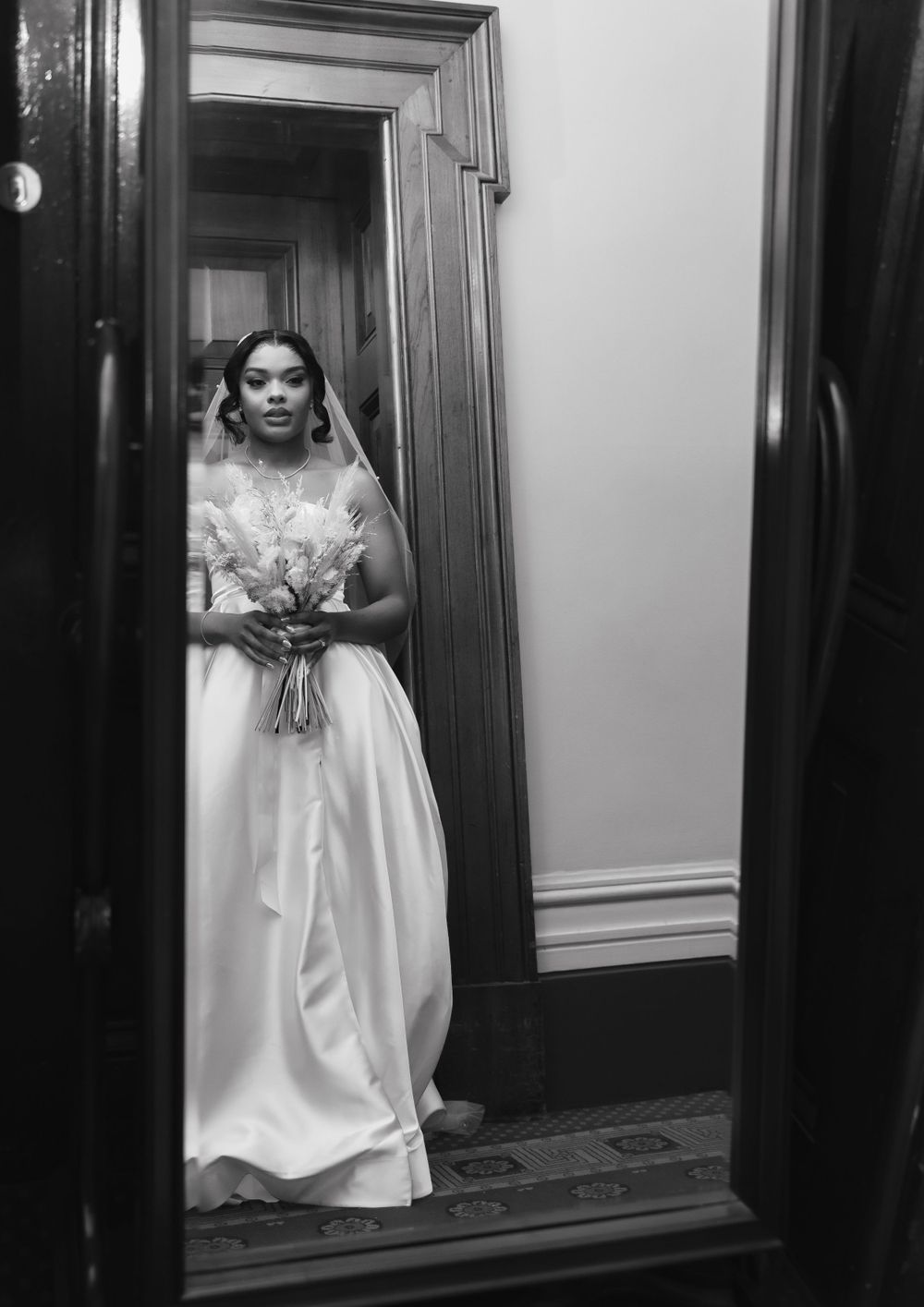 A black and white photograph of a bride in a flowing white gown and veil holding a bouquet, reflected in a mirror.