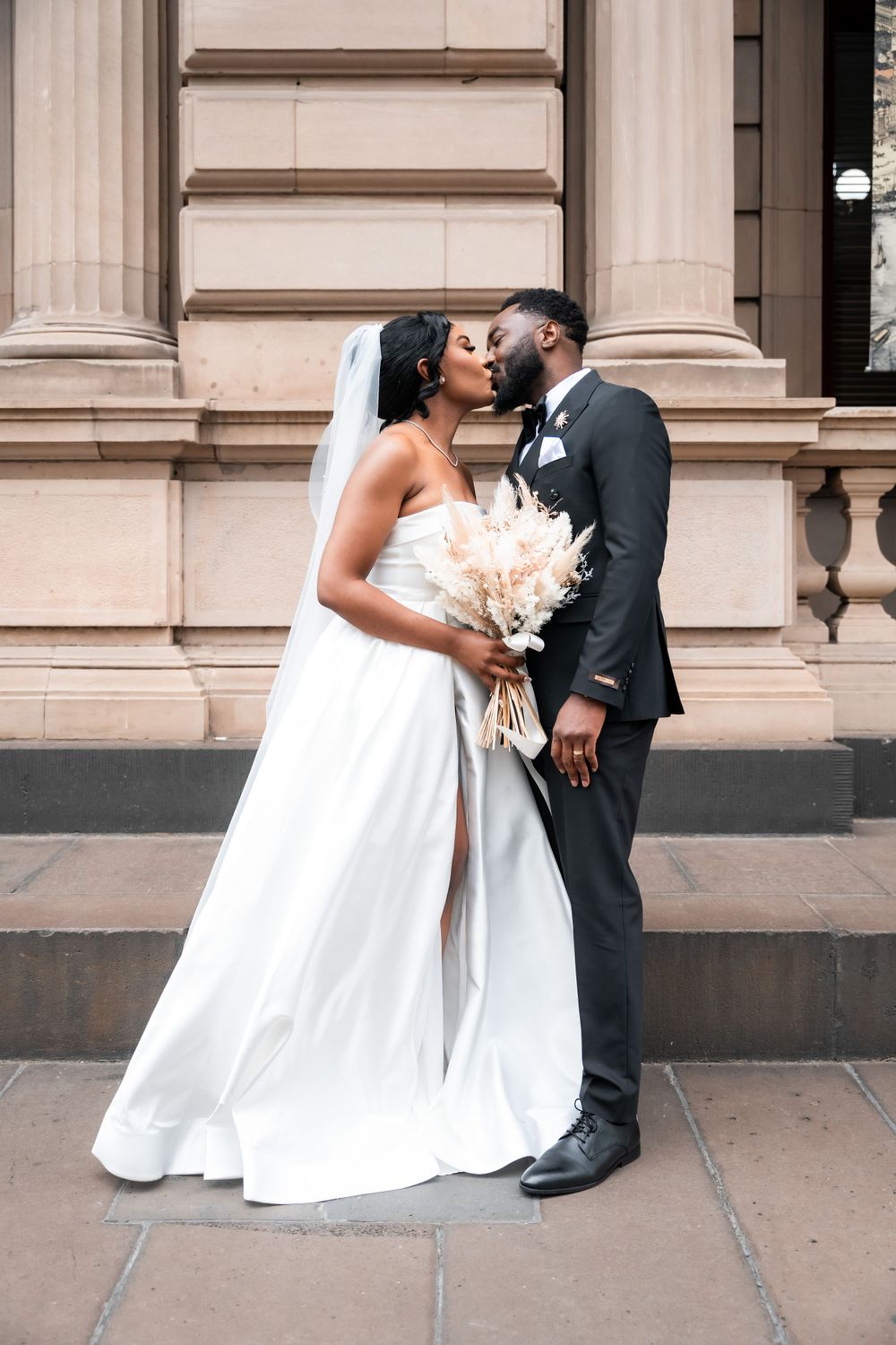 A romantic wedding couple shares a kiss on marble steps while the bride holds a delicate bouquet of white flowers.