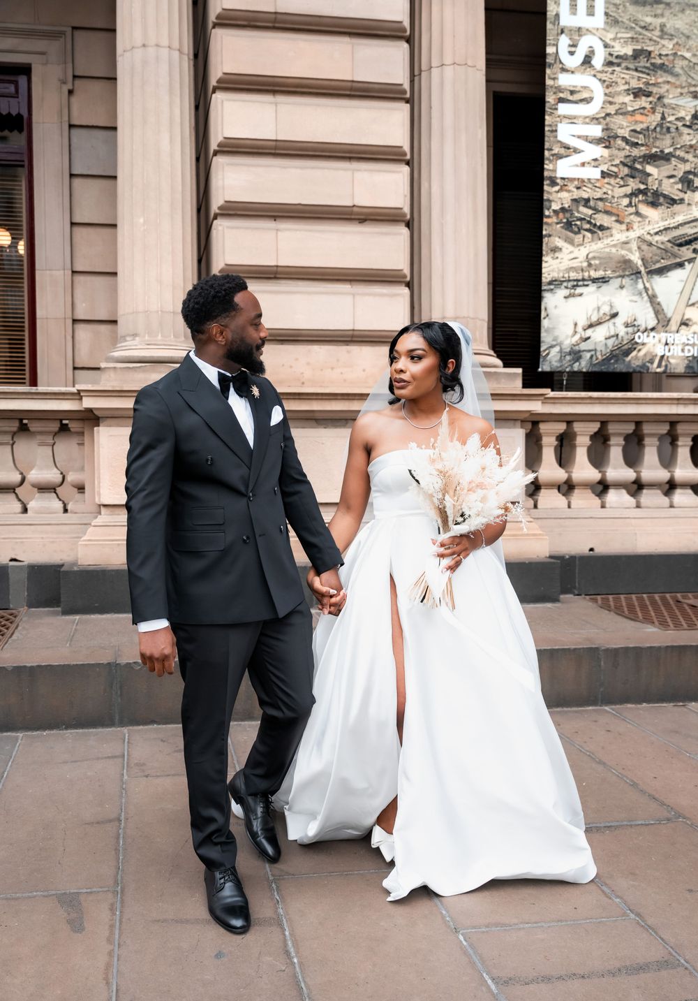 A couple in wedding attire hold hands on a grand stone balcony in front of an ornate building exterior.