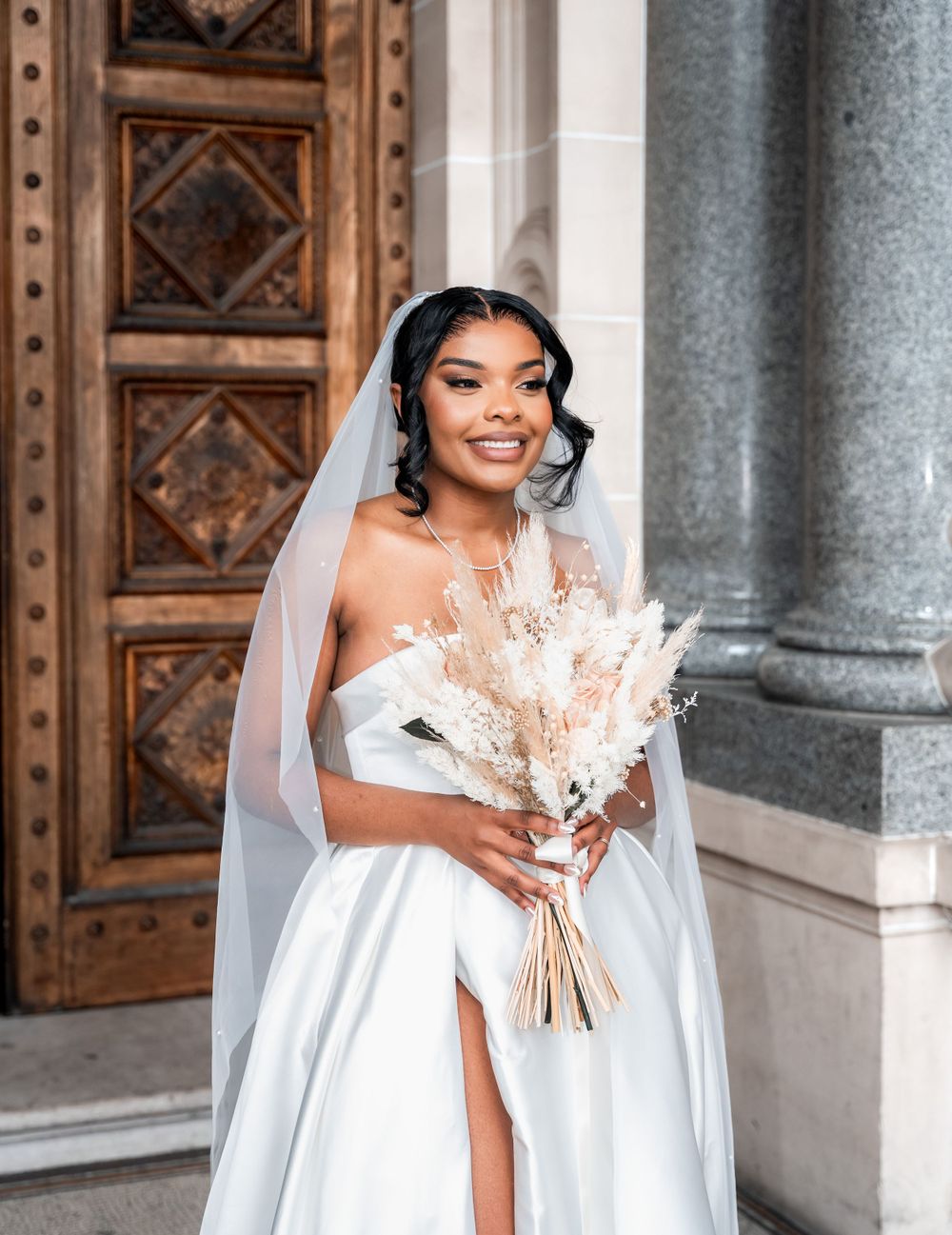 Elegant bride in a flowing white gown with thigh slit holds a dried pampas grass bouquet near ornate wooden doors and marble columns.