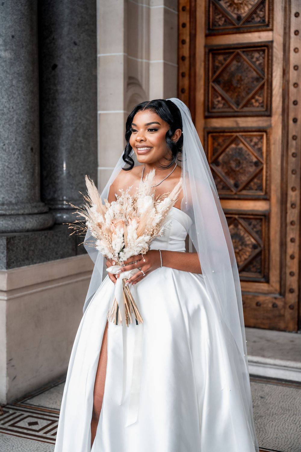 Beautiful bride in elegant white off-shoulder ball gown holds dried floral bouquet while standing by ornate wooden door.