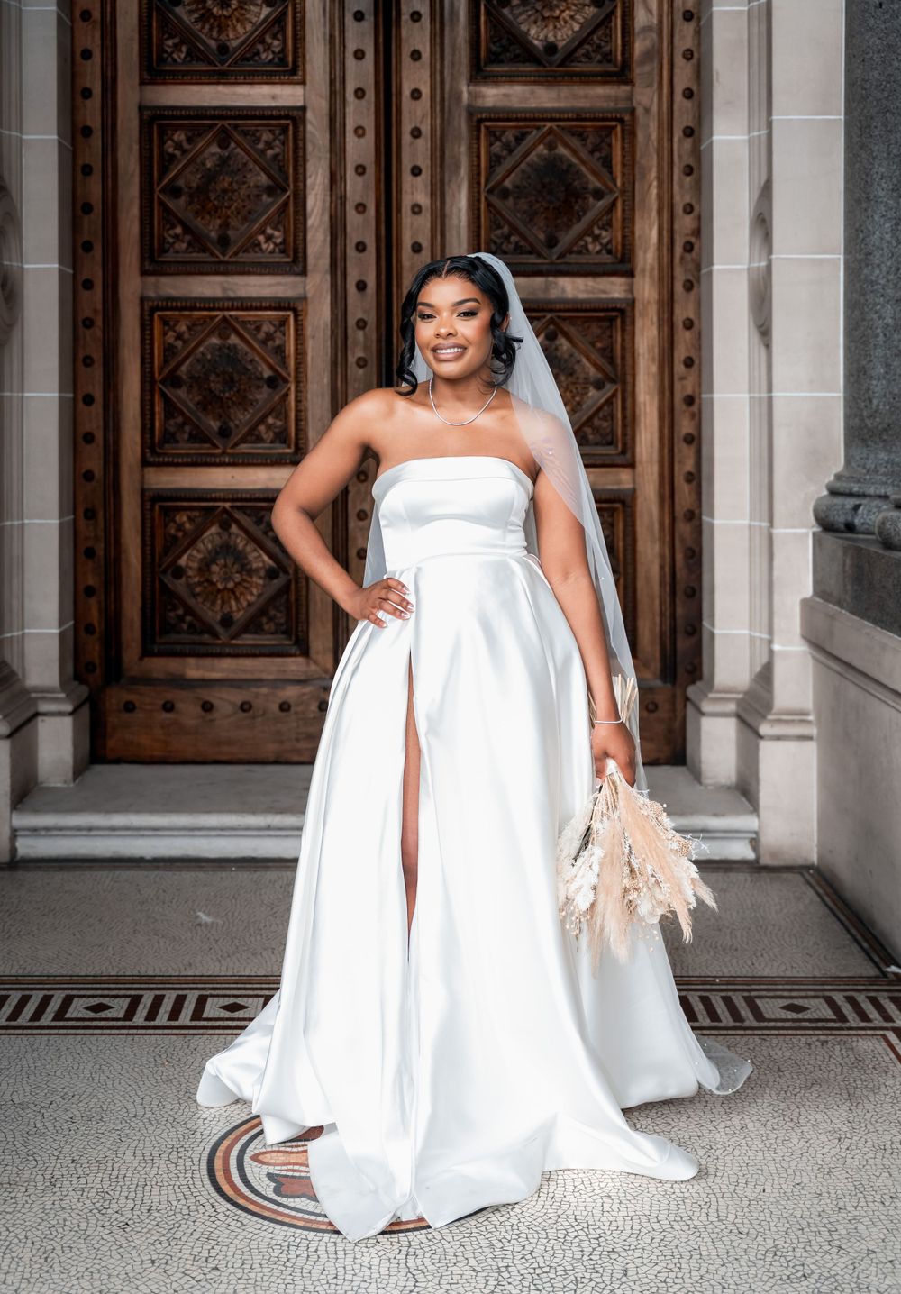 An elegant strapless white wedding dress with a high slit and cathedral veil stands in front of ornate wooden doors.