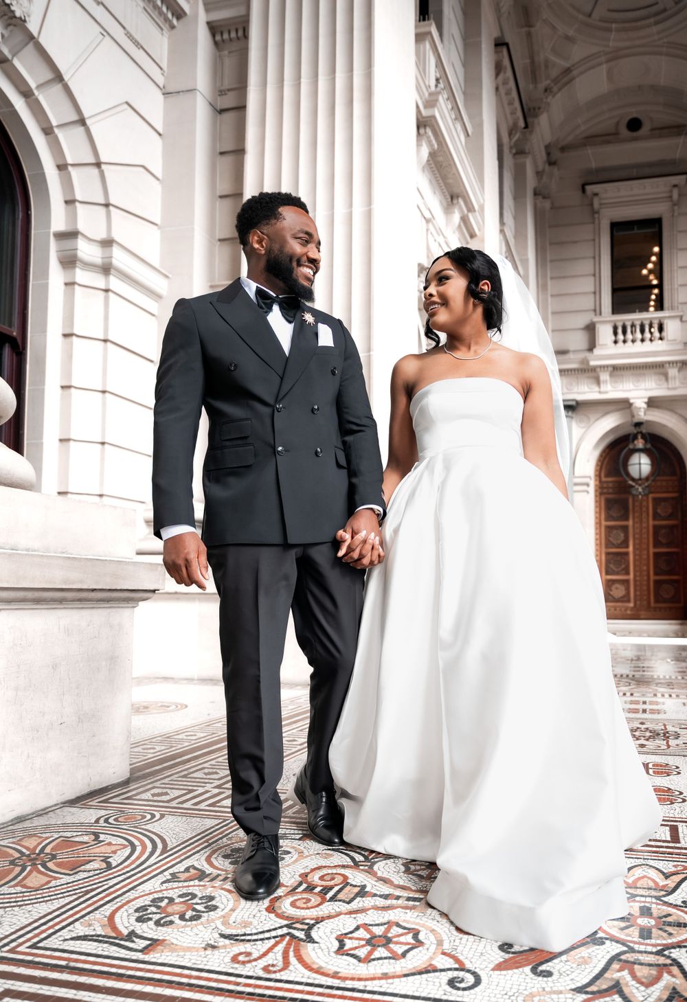 A couple in elegant wedding attire hold hands while standing on ornate marble flooring in front of grand columns.