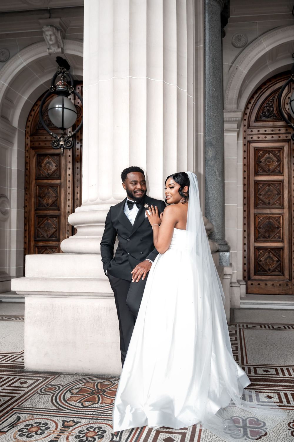 A bride in a white gown and groom in dark suit pose elegantly against classical columns with ornate wooden doors.