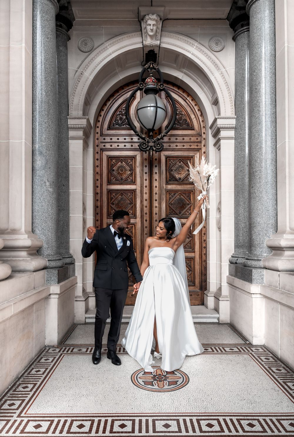 Newlywed couple celebrates joyfully under ornate stone archway with decorative door and mosaic floor.