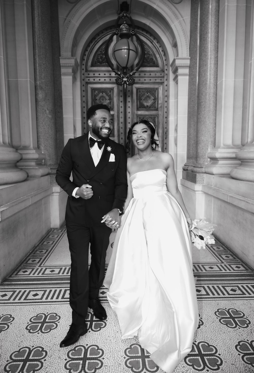 Black and white wedding photo showing newlyweds sharing a joyful moment in an ornate hallway with arched doorway and tiled floor.