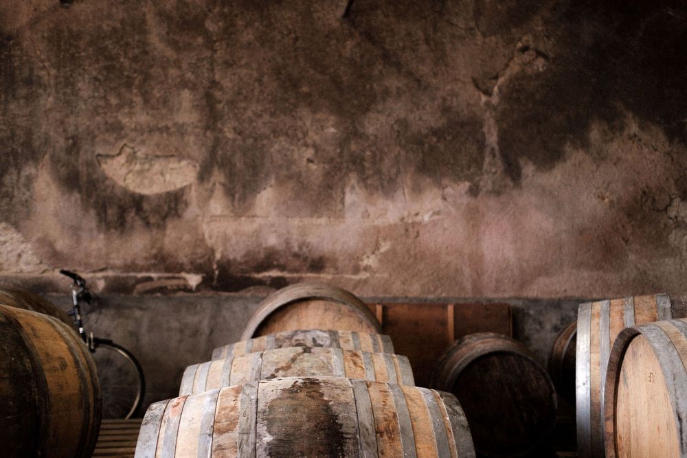 Aged wooden wine barrels stored against weathered stone walls in a rustic cellar.