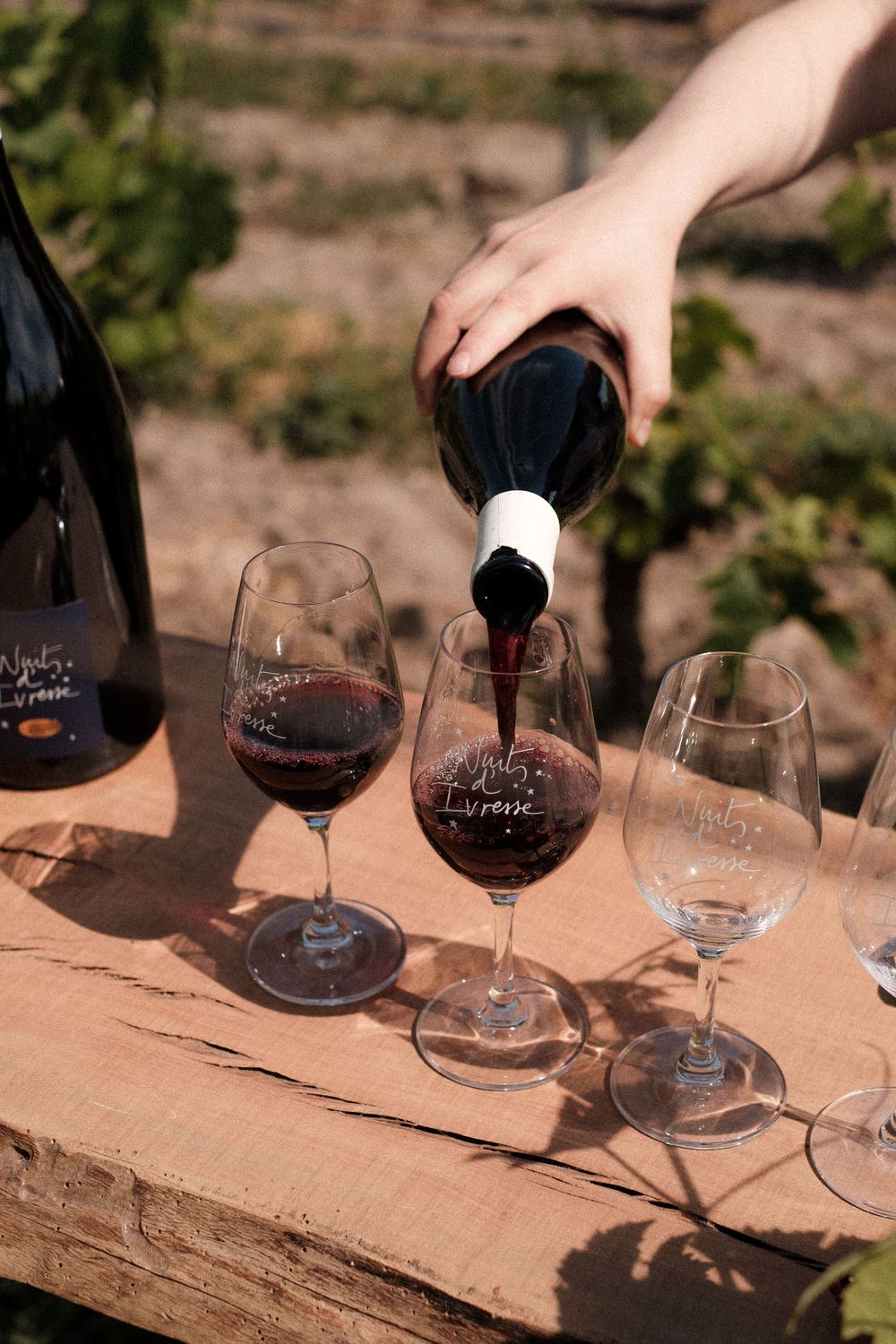 Hand pouring red wine into crystal glasses for tasting on an outdoor wooden surface.